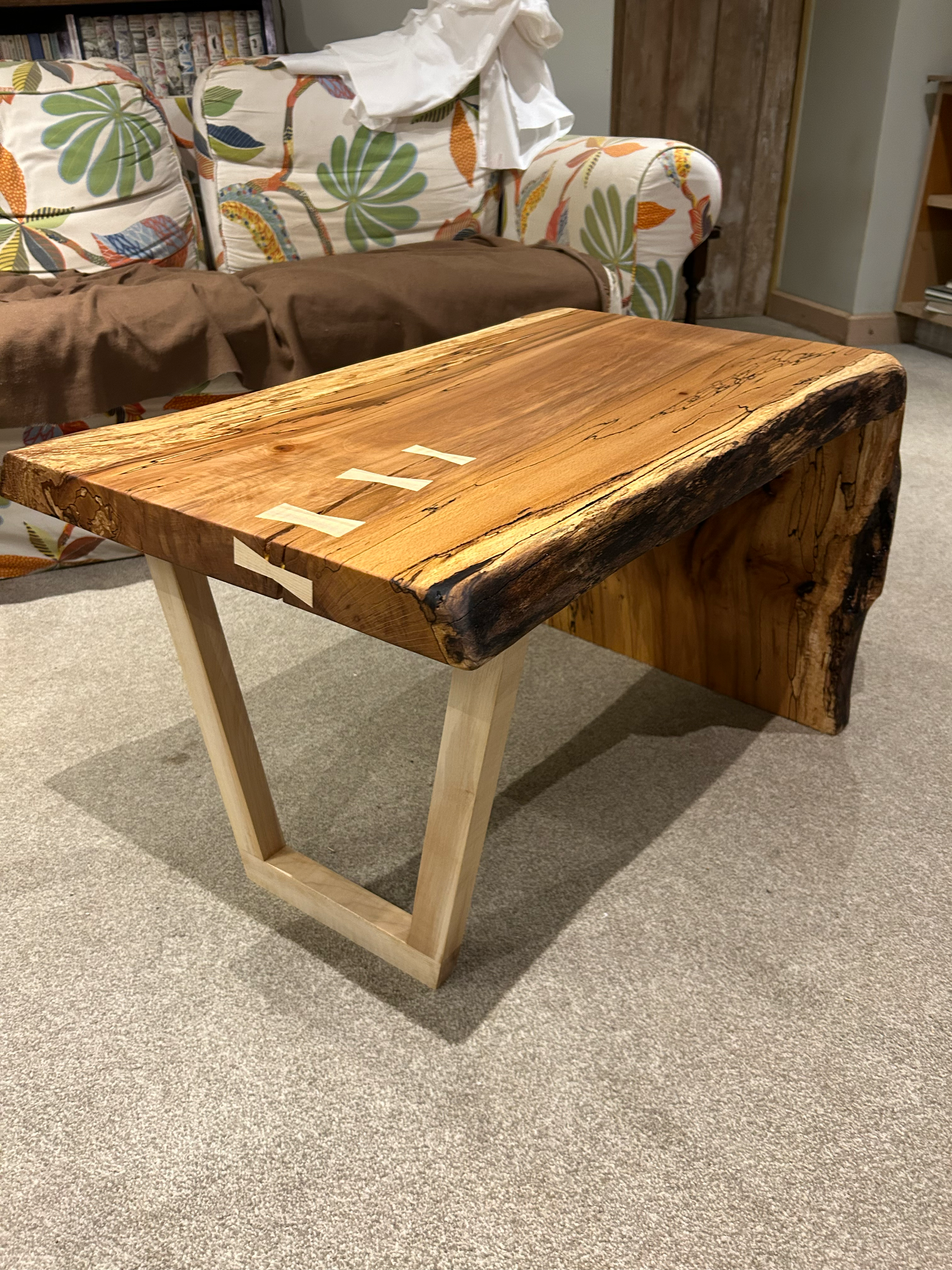 A rustic wooden table with a live edge, supported by a modern, light-colored wooden base, in a living room setting.