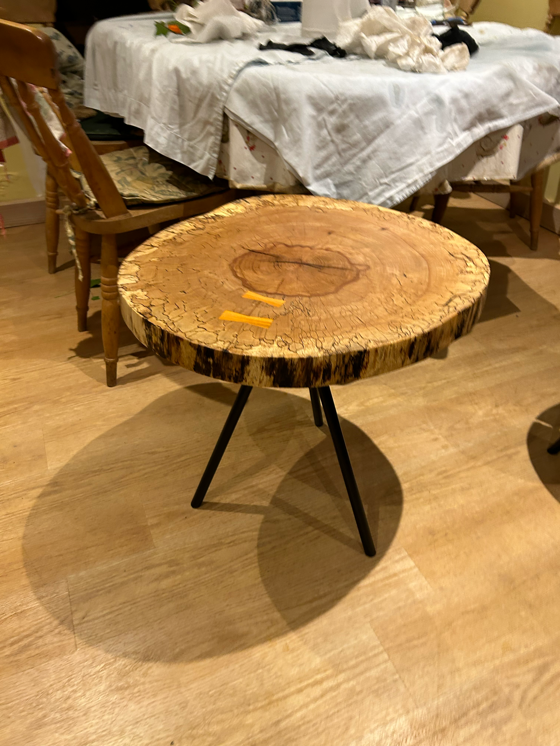 A rustic wooden coffee table with a thick, round slab of wood top and black metal legs, placed on a wooden floor in a room with a table covered in a white cloth and chairs in the background.