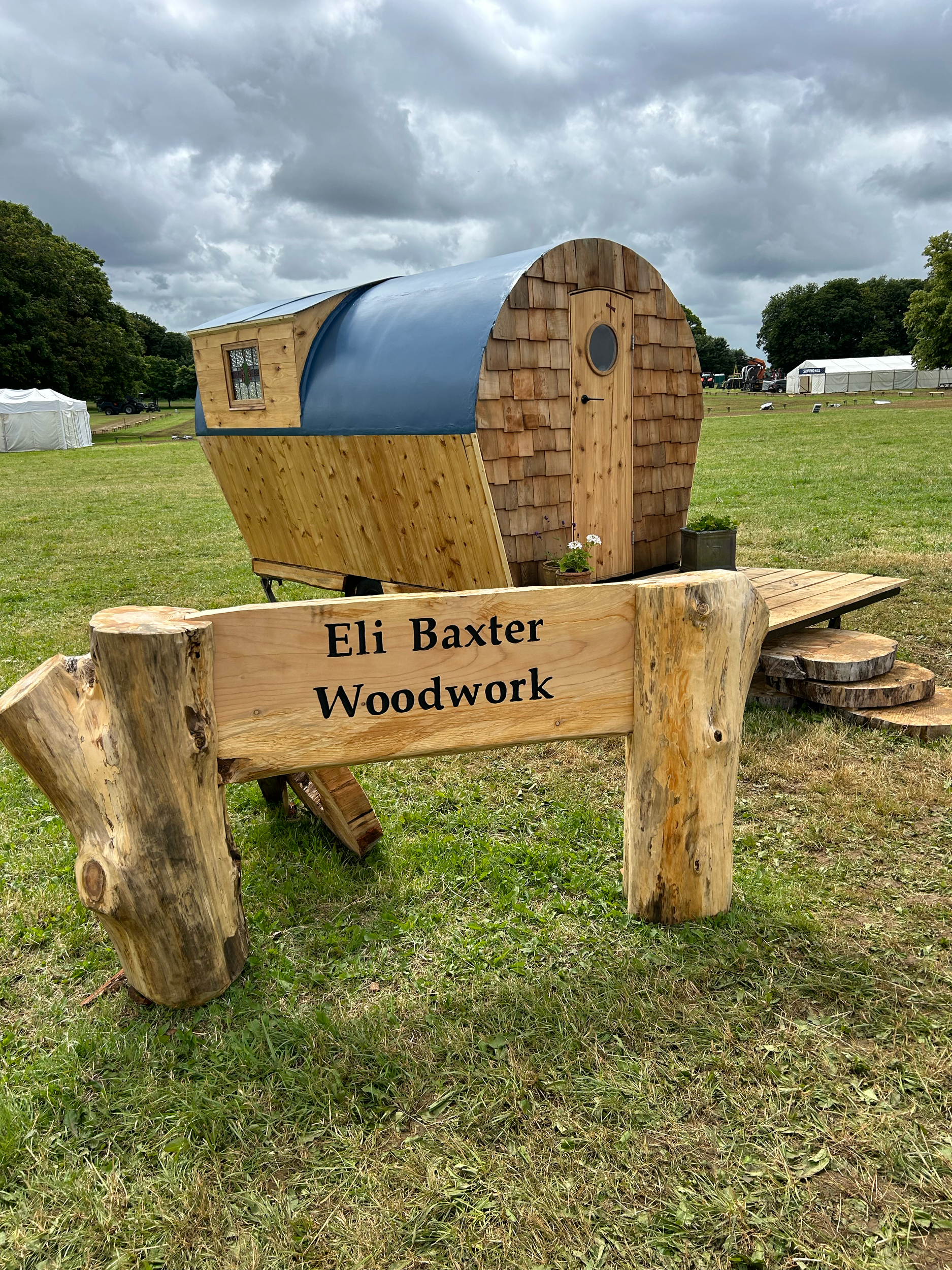 A small, wooden tiny house with a rounded door, small window, and shingled exterior on a grassy field. In front, there is a rustic sign saying 'Eli Baxter Woodwork' made from thick wood logs.