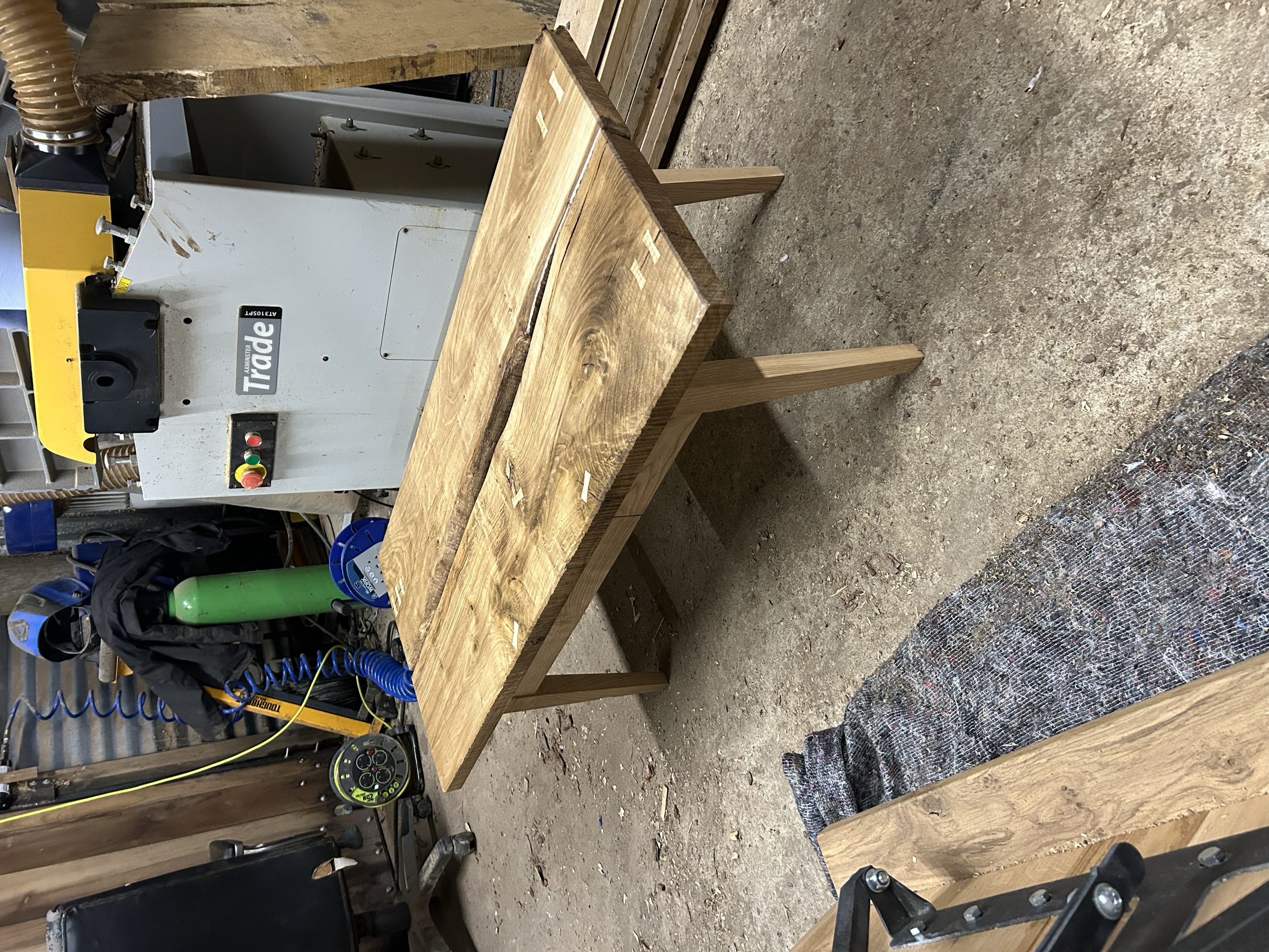 A wooden workbench in a woodworking shop with a large machine behind it and various tools and objects around.