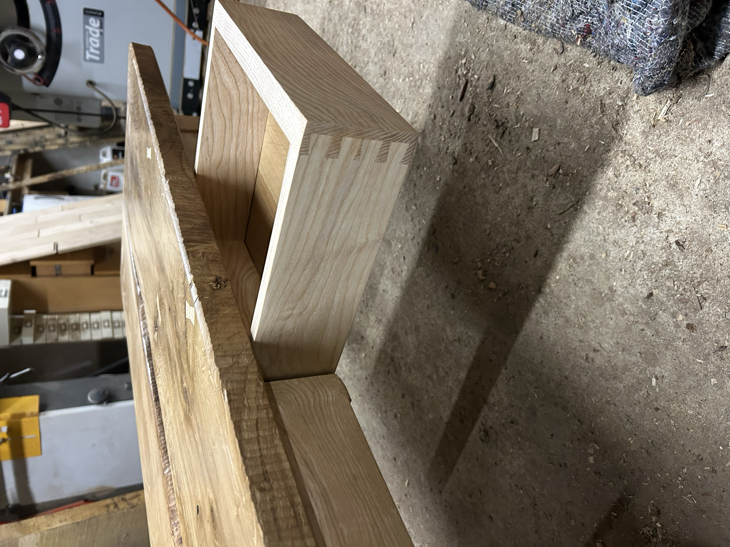 Unfinished wooden piece with a smooth top surface and a mortise cut into the side, placed on a dusty workshop floor next to a colorful woven cloth.