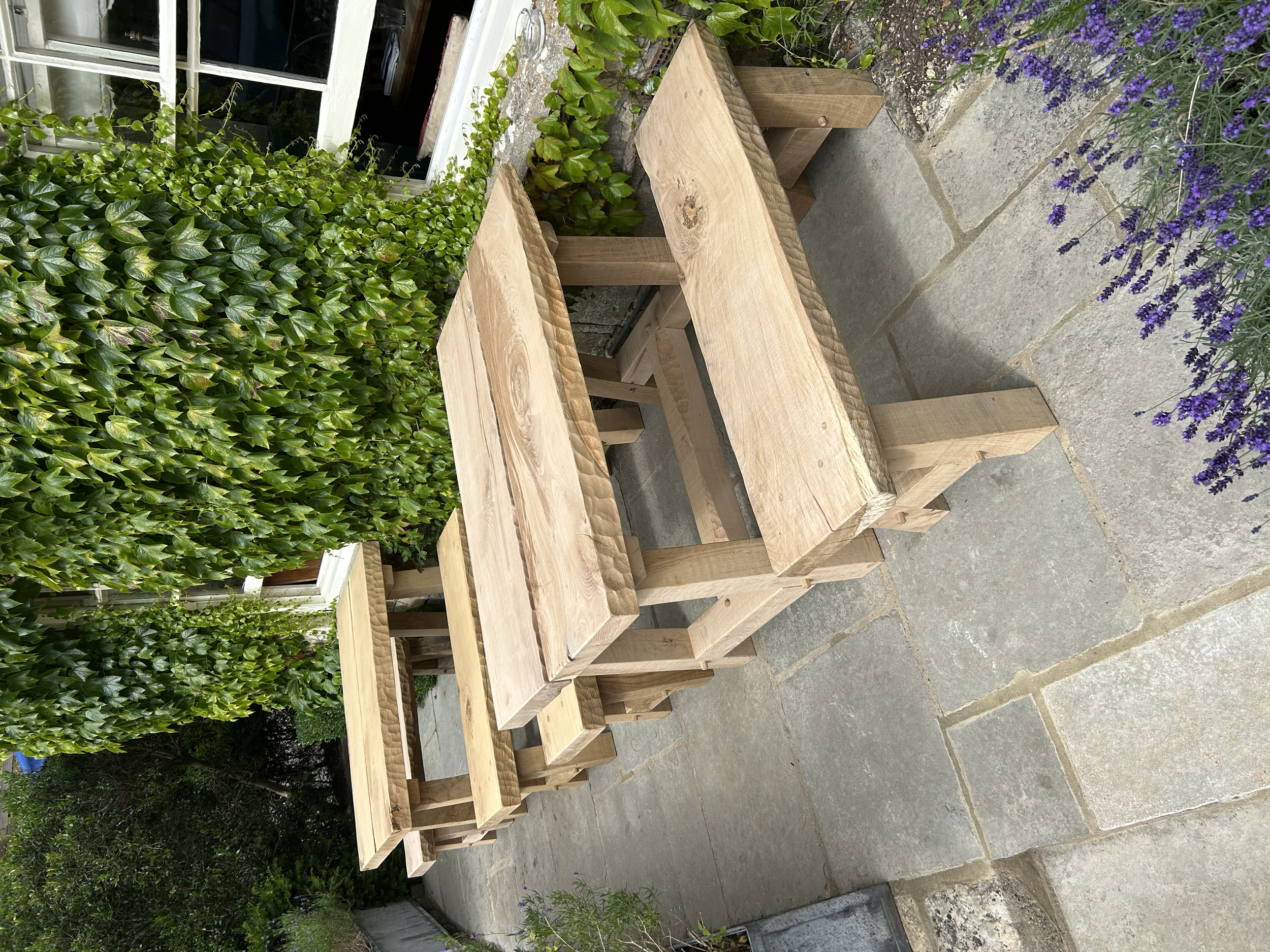 A wooden outdoor table with matching benches on a stone patio, with green hedge and purple flowers in the background.