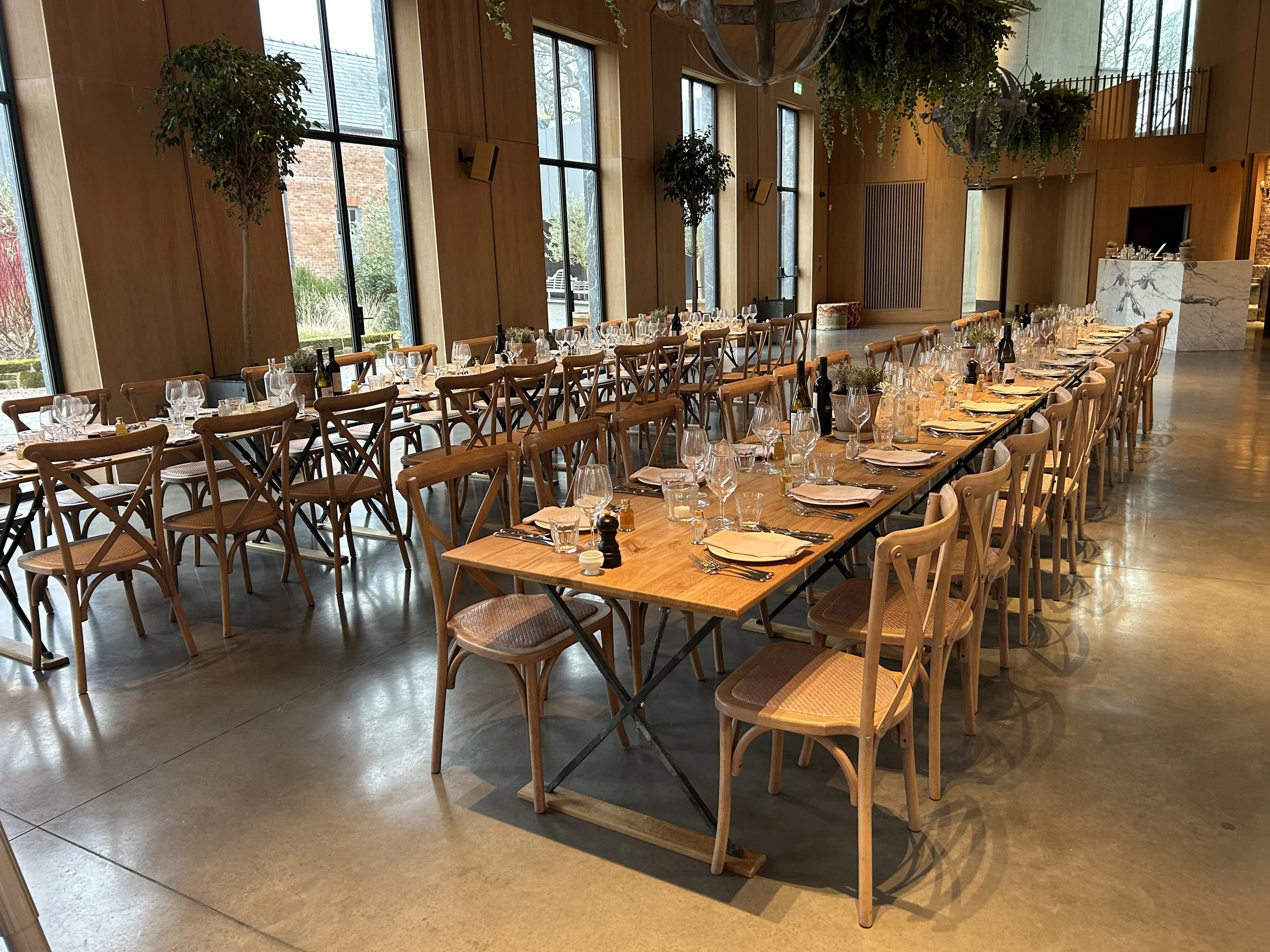 Empty banquet hall with long wooden tables set with wine glasses, plates, silverware, napkins, and small plants, featuring large windows, wooden walls, and hanging greenery.