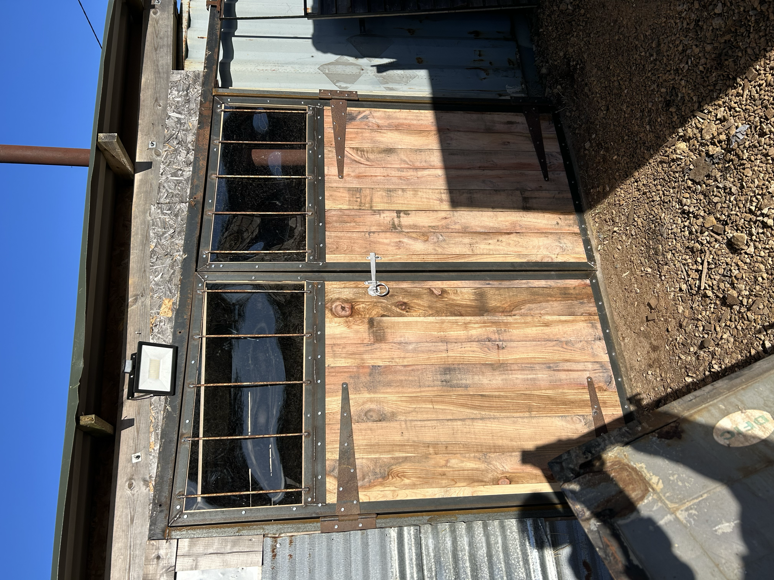 A barn-style door with metal hinges and a lock, two small barred windows, and a metal roof. The door is made of wooden planks and is part of a rustic shed or stall, with brown dirt ground in front and sunlight casting shadows.