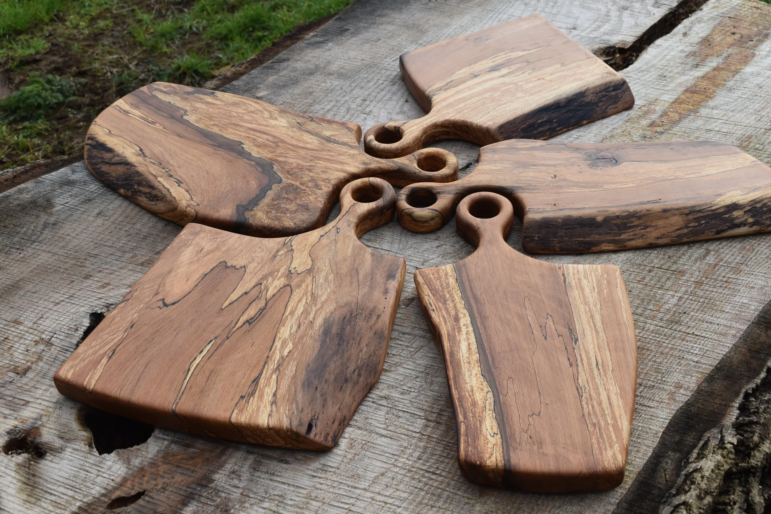 Four wooden cutting boards with rounded handles are arranged on a weathered wooden surface outdoors.