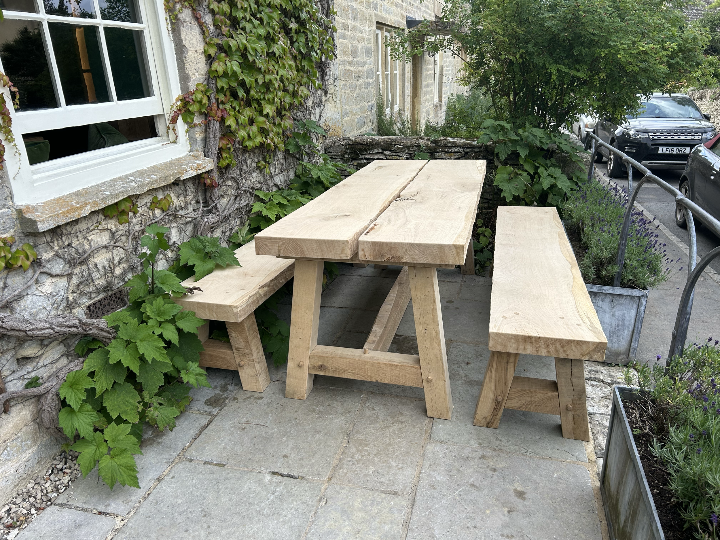 Outdoor patio scene with a wooden table and two benches, next to a stone house wall with ivy and window, surrounded by planters with green and purple plants, and parked cars on the street.