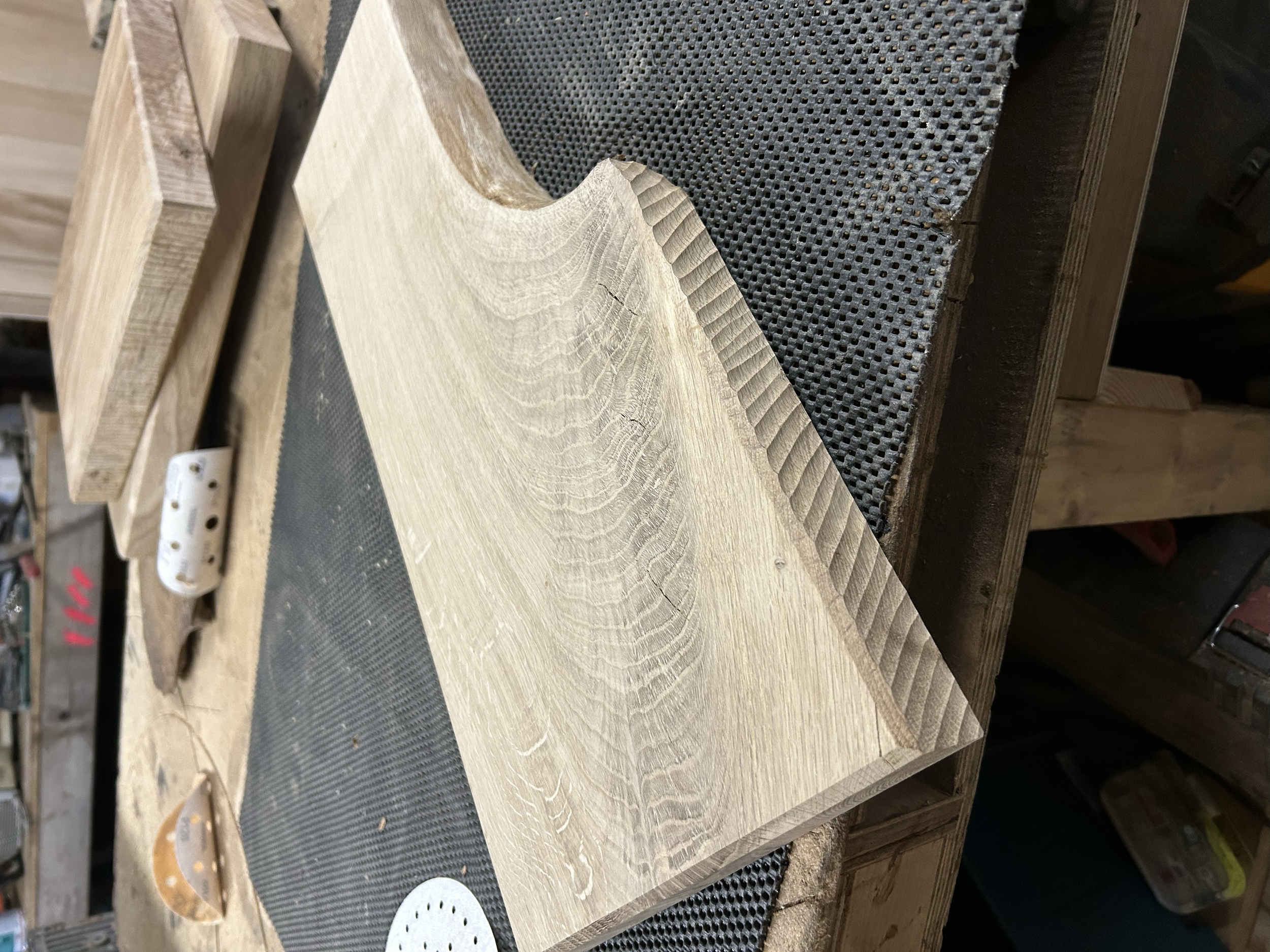 A piece of light-colored wood with a carved semi-circular notch on one side, resting on a black textured work surface in a woodworking shop.