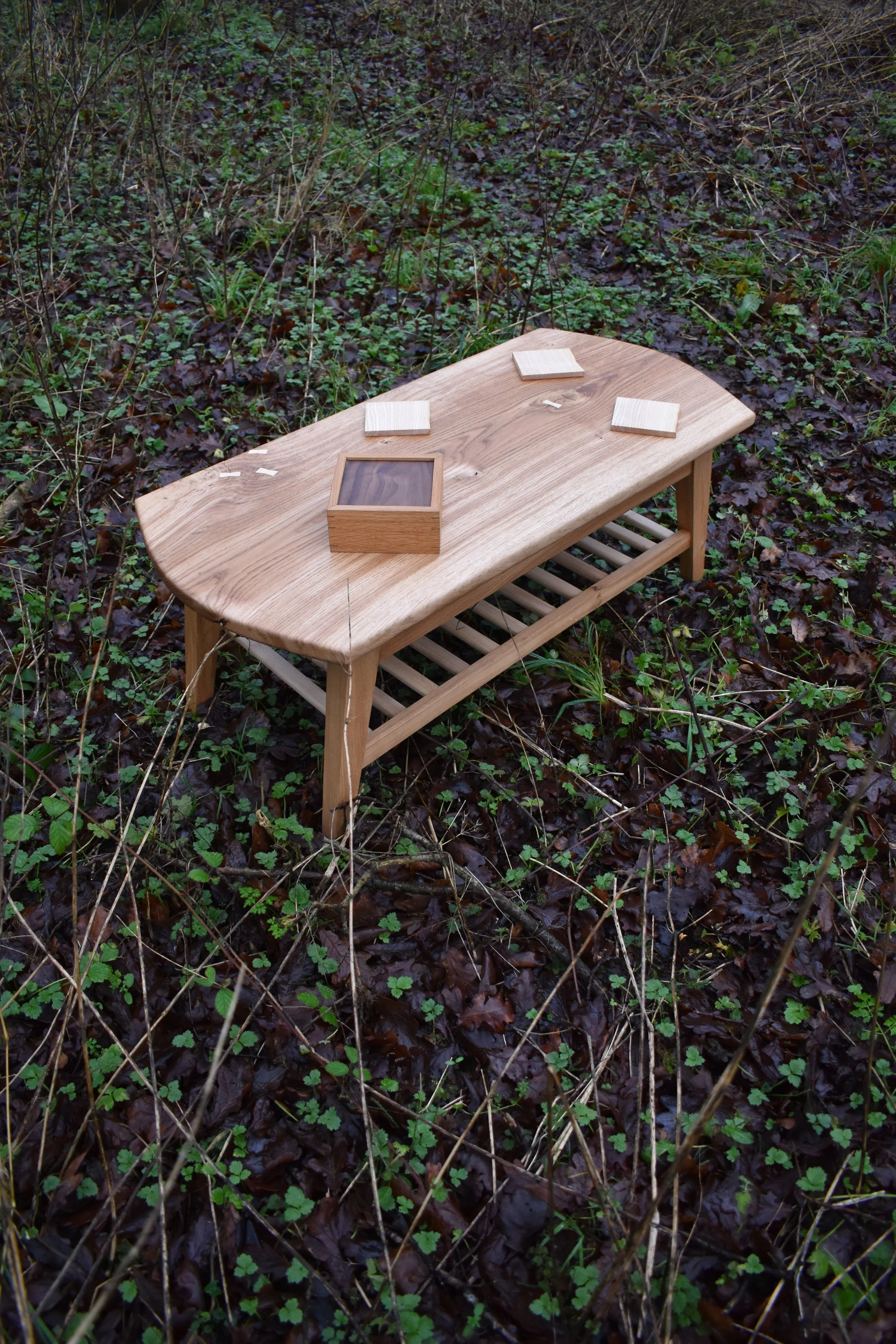 A wooden table outdoors on a patch of ground covered with wet leaves and small plants, with three square coasters and a small wooden box on top.