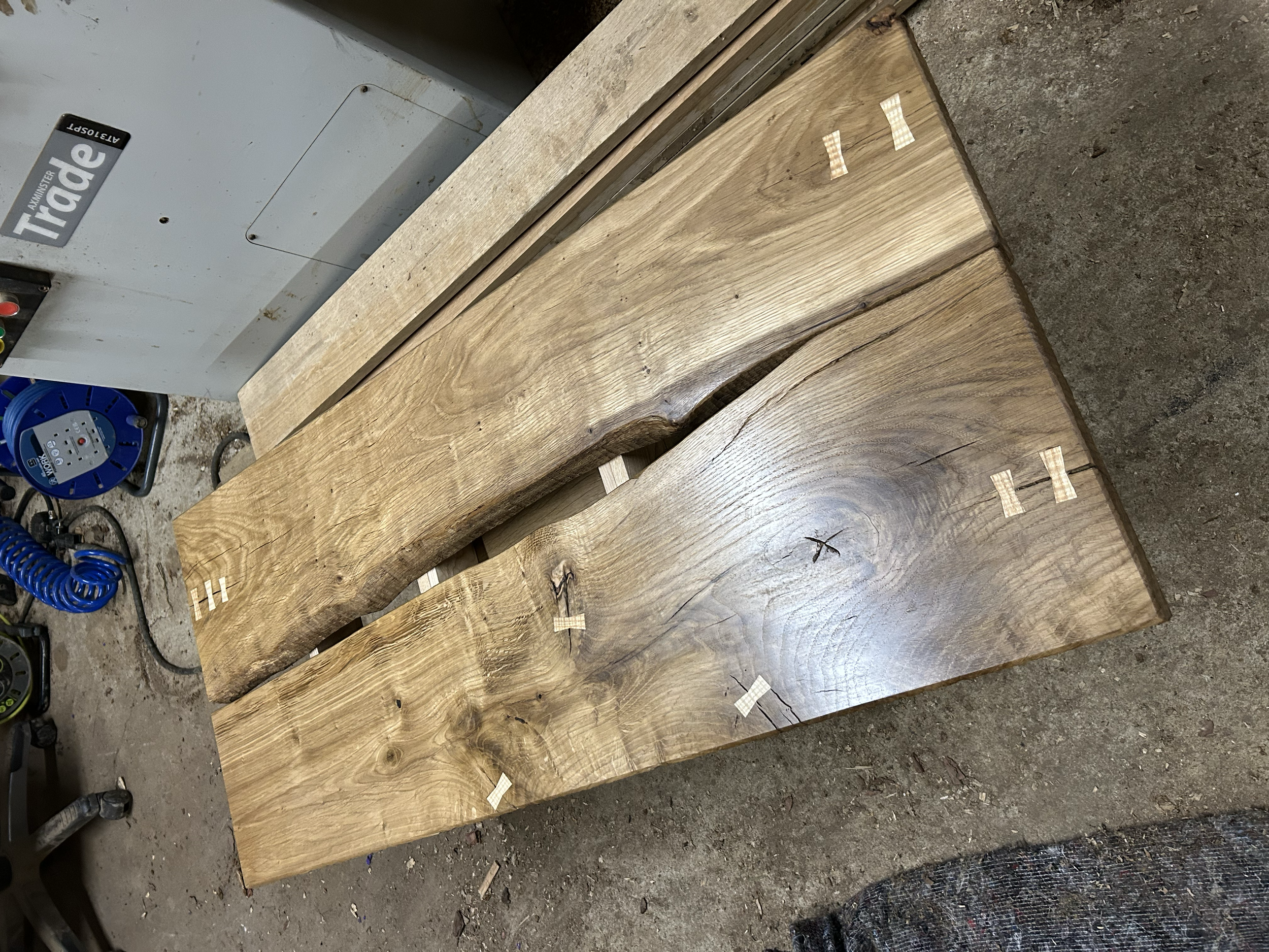 Two large wooden slabs with visible wood grain and knots, marked with small wood pieces, placed on a workbench in a workshop environment.