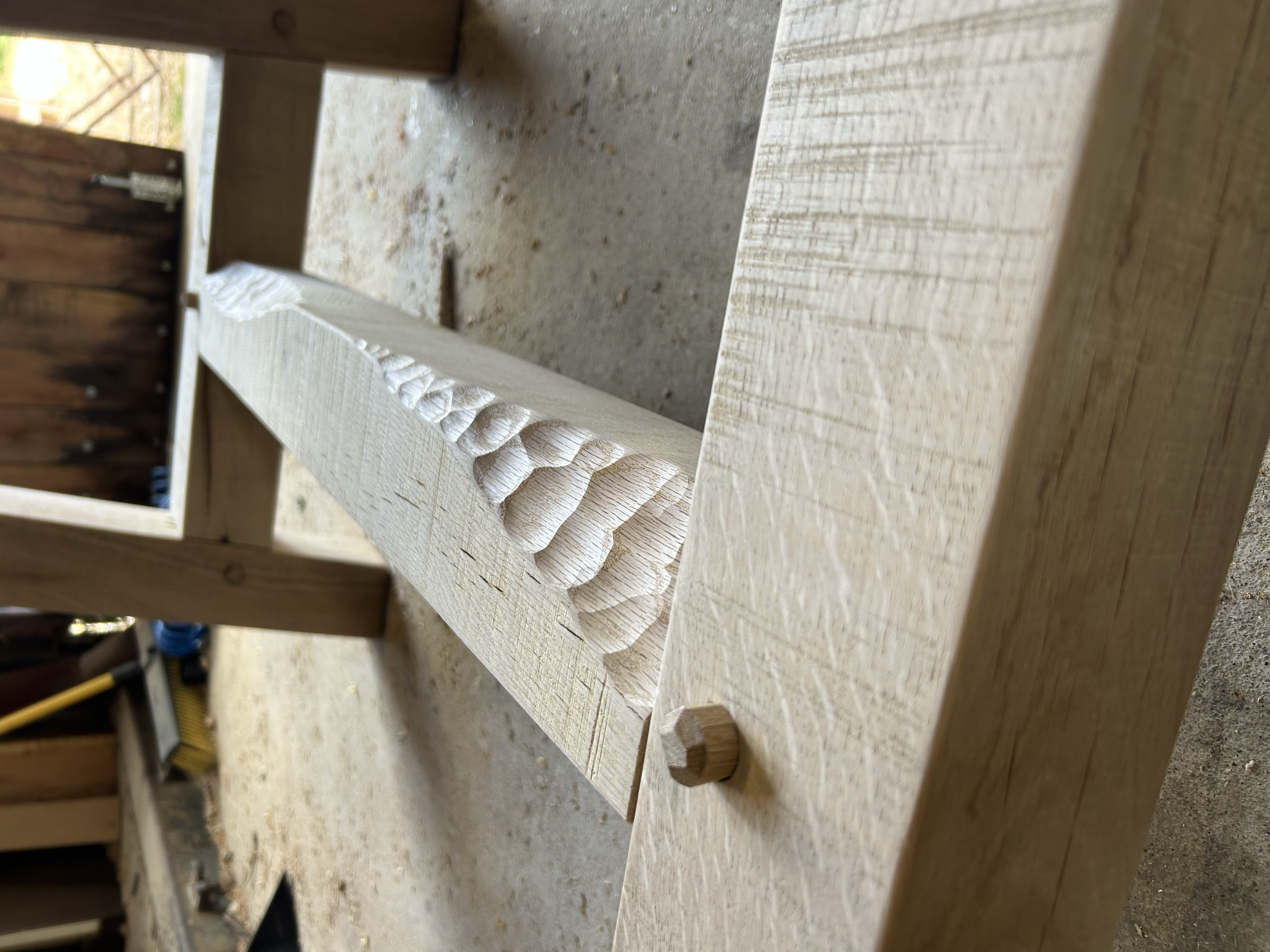 Close-up photo of a woodworking project on a workbench, showing a piece of wood with a carved pattern and a small wooden peg.