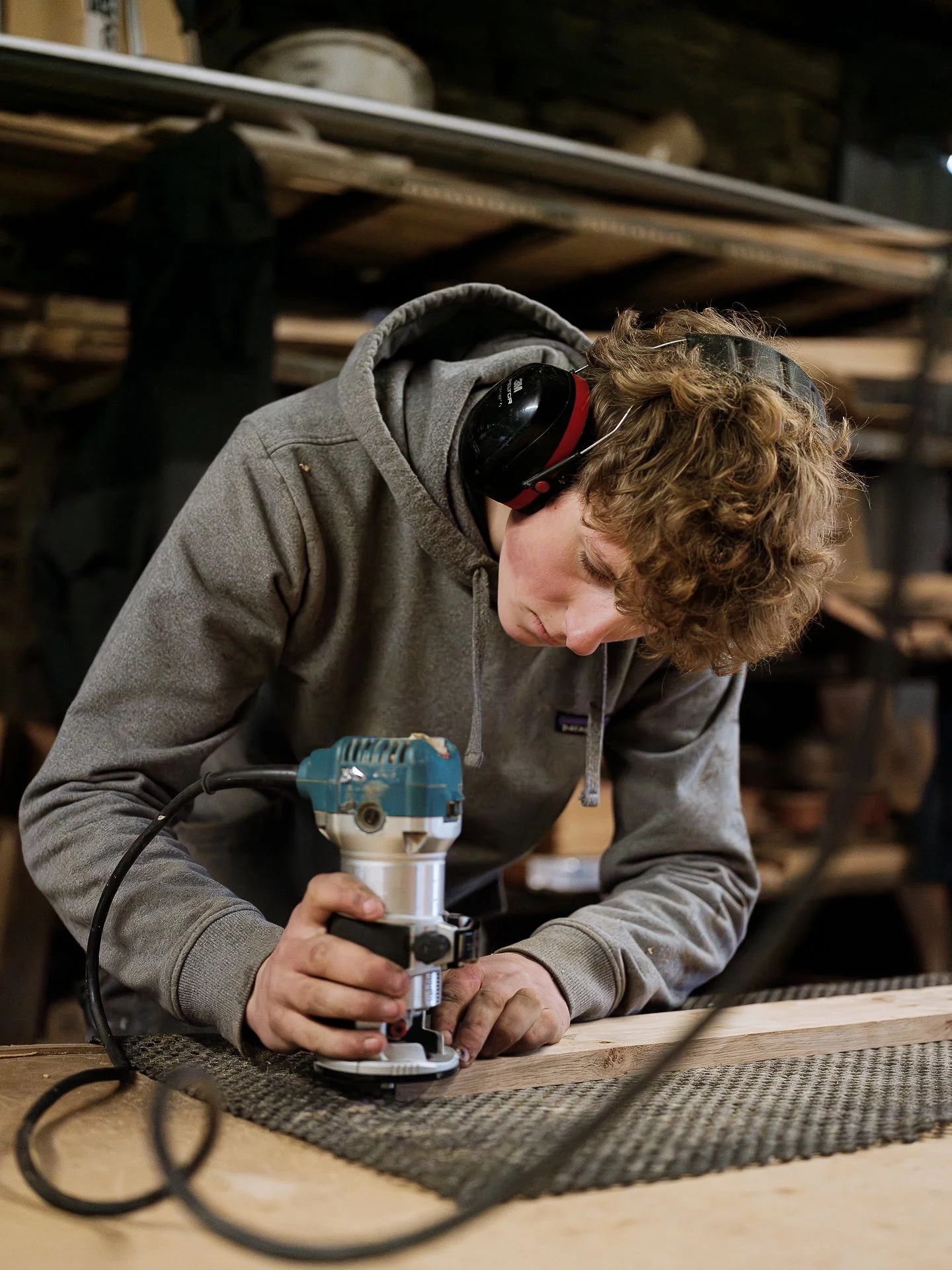 Young man working with a router on a piece of wood in a woodworking shop, wearing gray hoodie and safety ear protection.