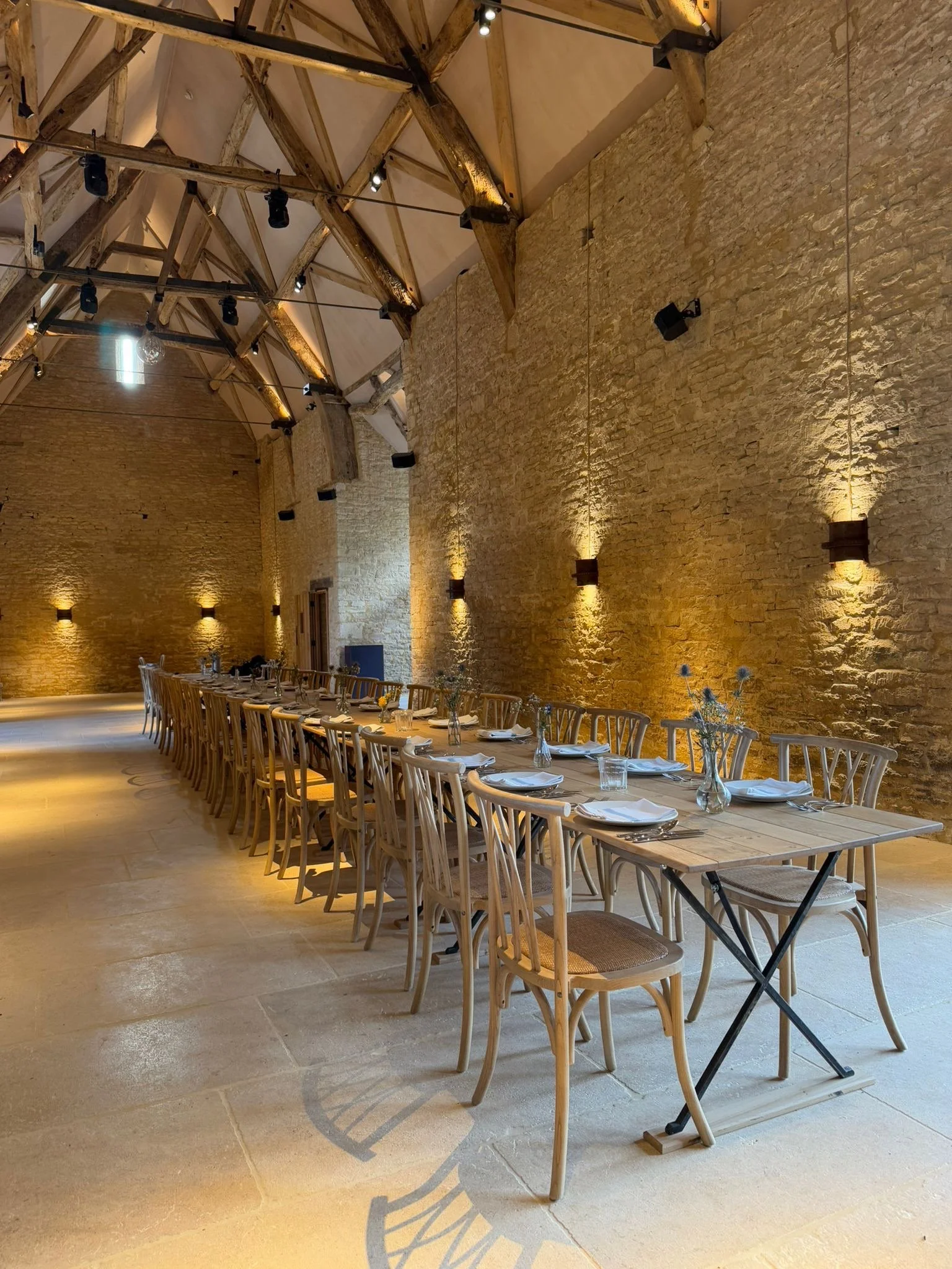 Long wooden dining table set with white plates, glasses, and small vases with flowers in a rustic, brick-walled event space with wooden ceiling beams and warm lighting.