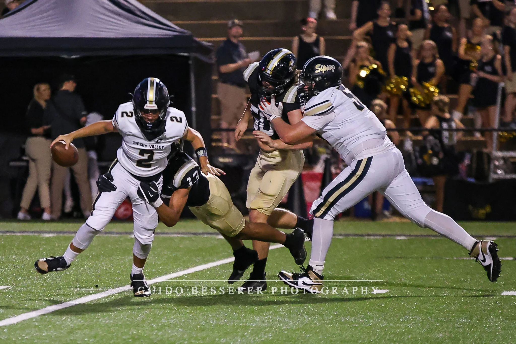 Football players from Nipsey Valley team in action during a game, with one player running with the ball, while others attempt to block or tackle, on a football field at night.