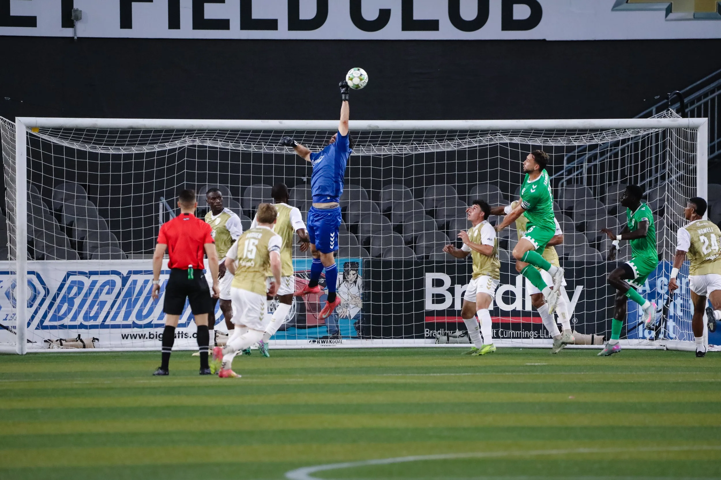 A soccer match with players jumping for the ball in front of the goal. The goalkeeper in green is attempting to block the shot, while the other players watch and prepare for the next move. An official in a red shirt is also present on the field.