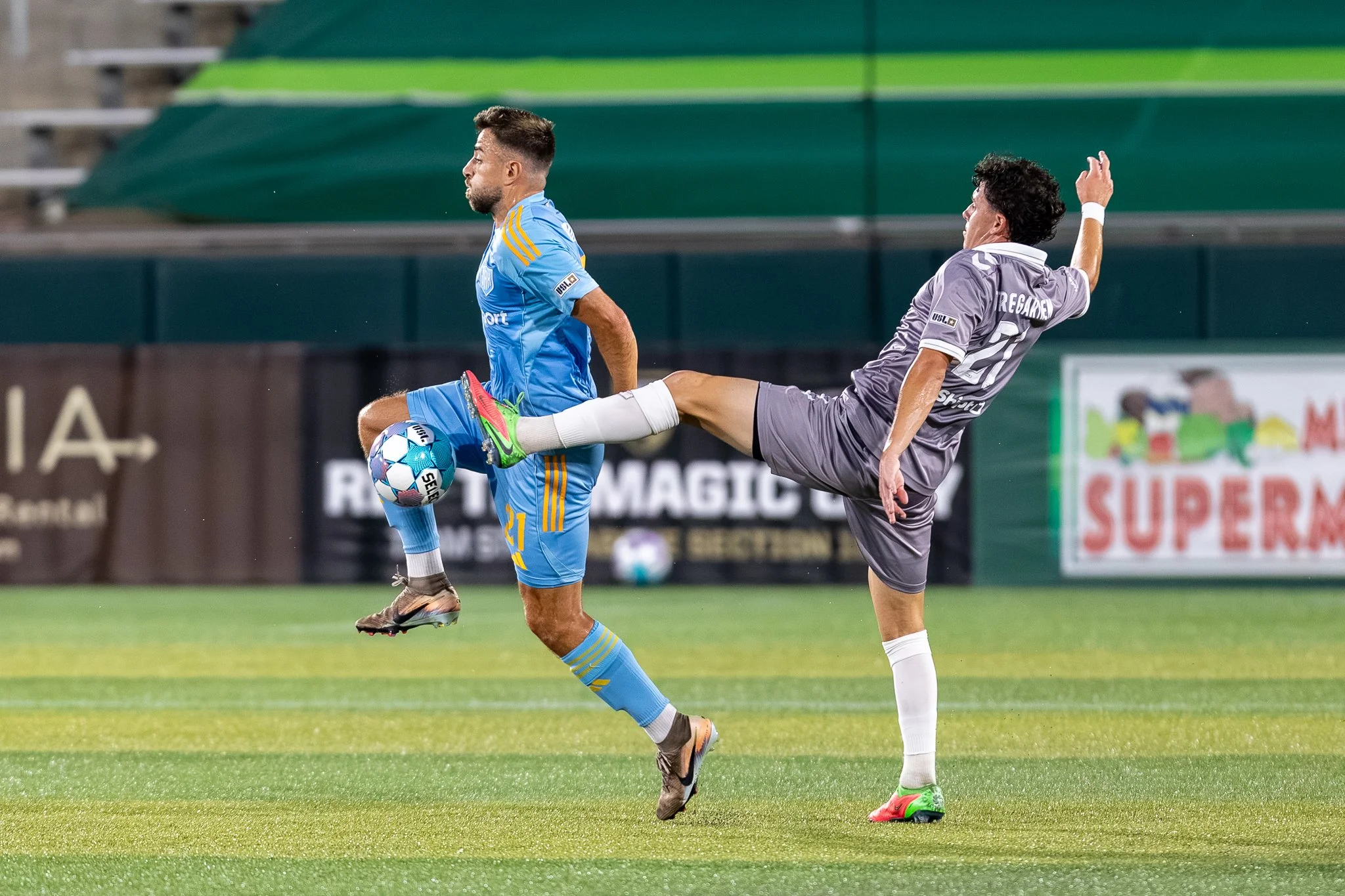 Two soccer players compete for the ball during a match on a green field, with one player wearing a light blue jersey and the other in a gray jersey, with stadium seating in the background.