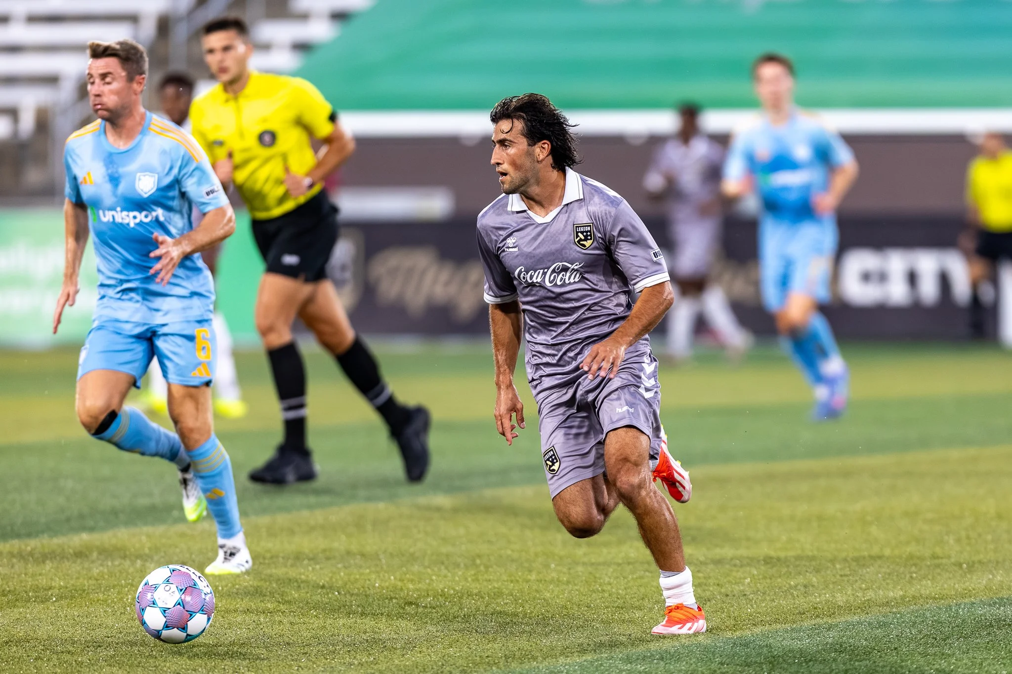 A soccer player in a gray jersey and orange shoes runs on the field near the ball during a match, with other players and a referee in the background.
