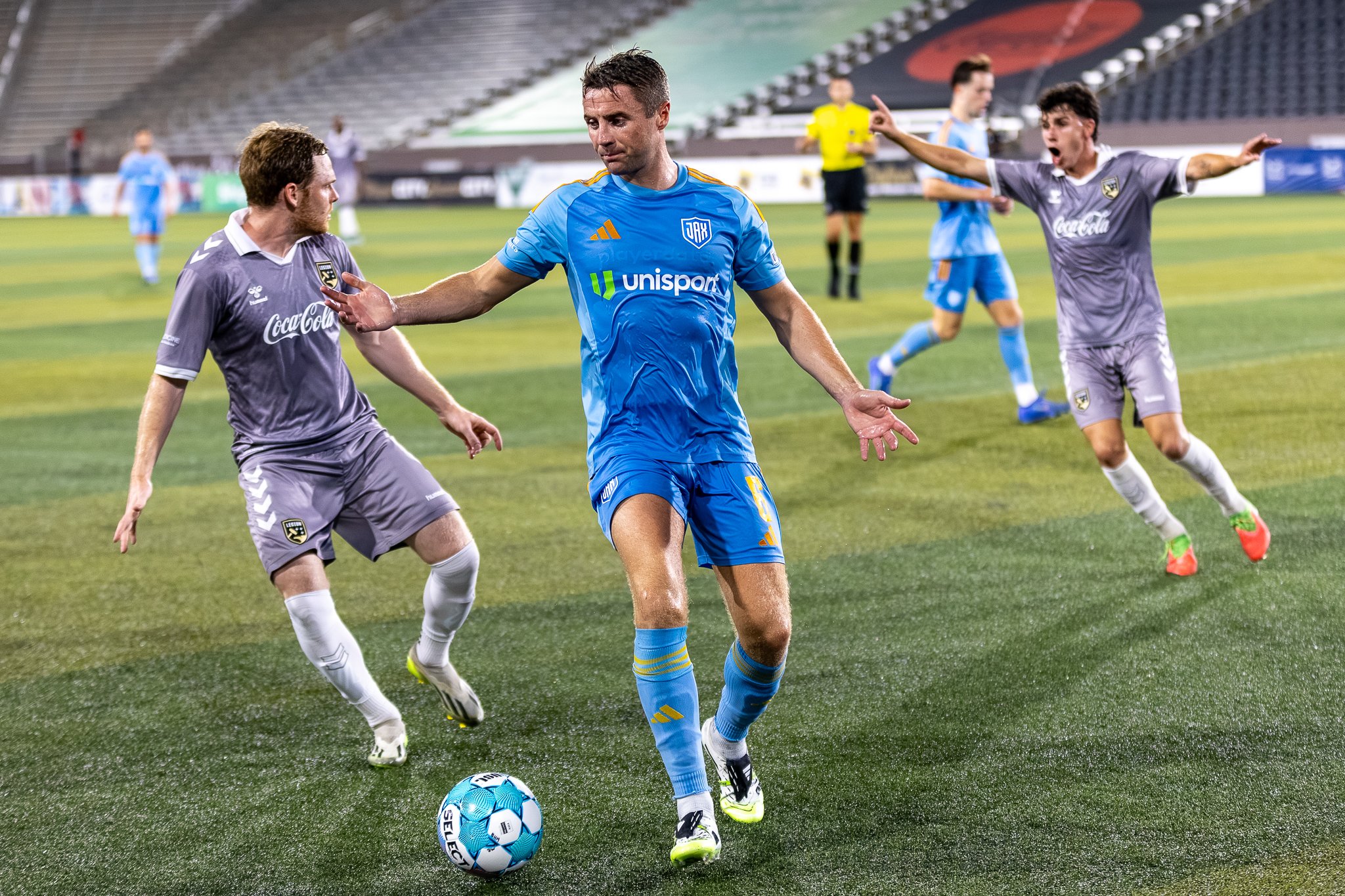 Soccer match with players in blue and gray uniforms on a green field, with a player in blue controlling the ball as opponents reach out for it.