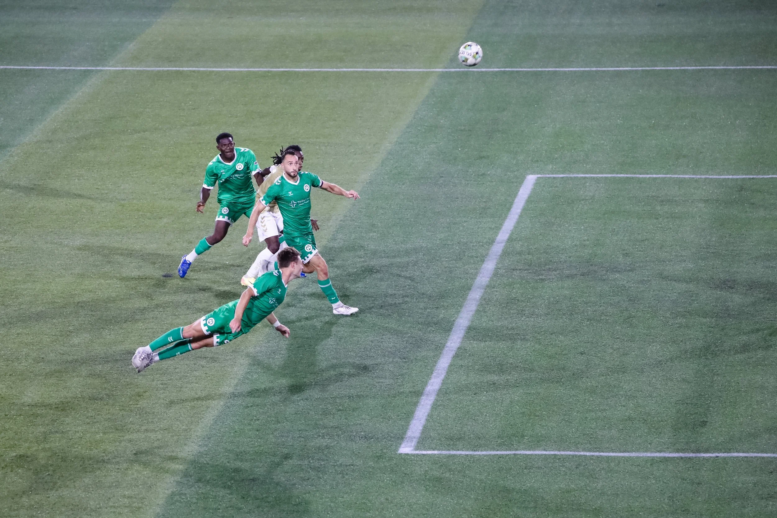 Four soccer players in green uniforms are on the field, with one diving to attempt to kick the ball, while the others are running or watching. The ball is mid-air near the goal area on a green field.