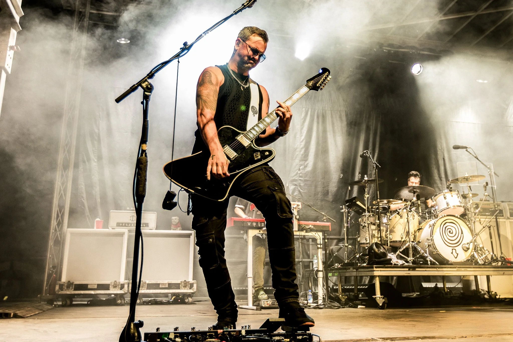 Musician playing an electric guitar on stage during a concert with fog and stage lights, with a drummer in the background.