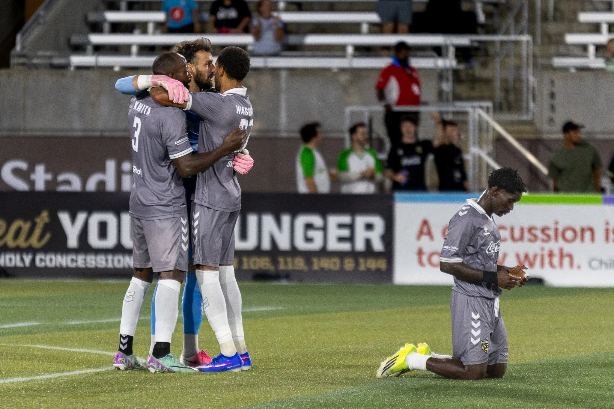Three soccer players wearing gray uniforms hugging each other, one standing behind the others, in a stadium field. Another player in gray kneeling on the grass, looking down while holding a phone. The background shows advertising boards, other people