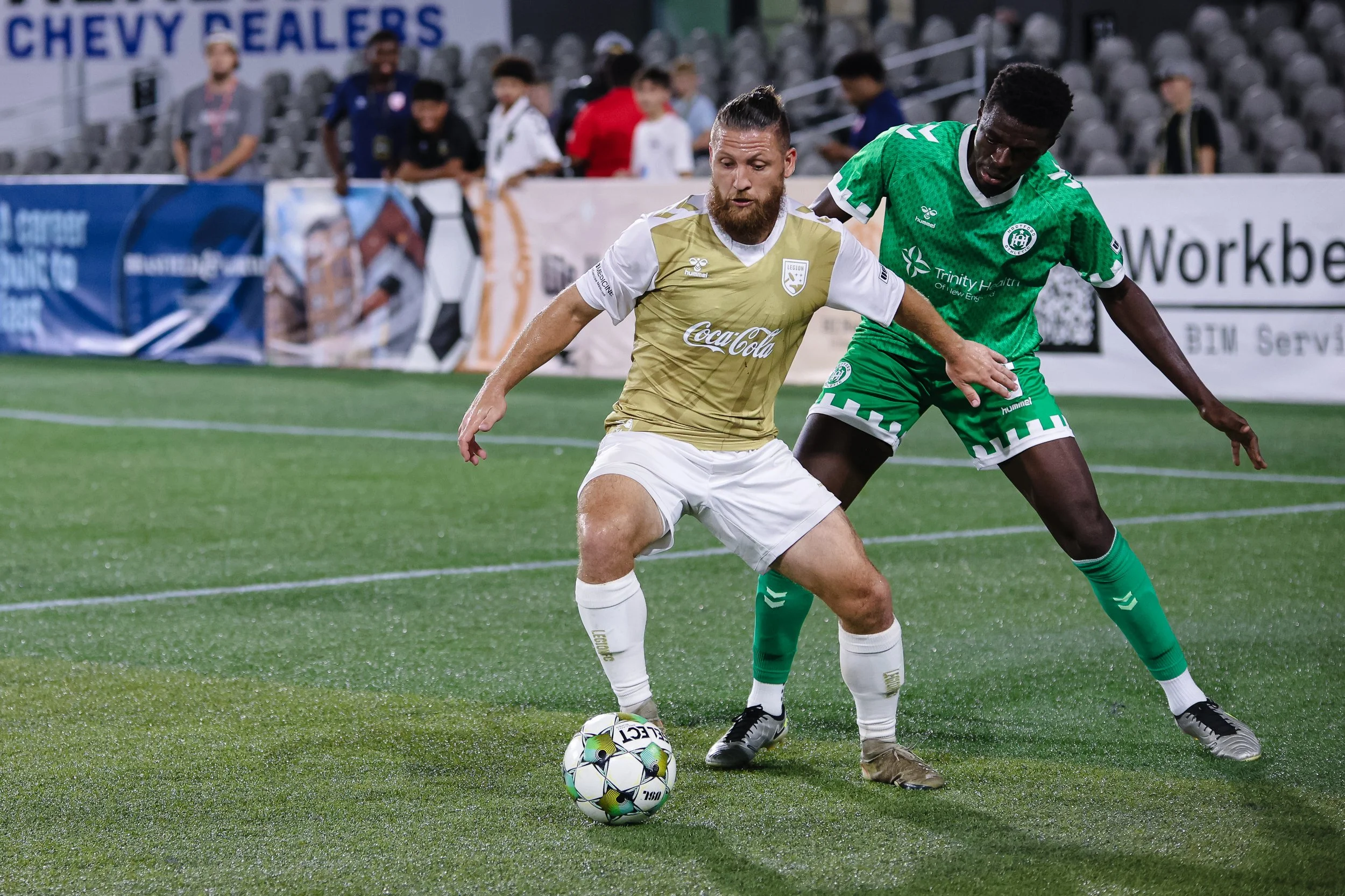 Two soccer players contest a ball during a match. One wears a gold and white uniform with a Coca-Cola logo, the other in a green kit with white accents. They compete on a green field with fans and advertising banners in the background.