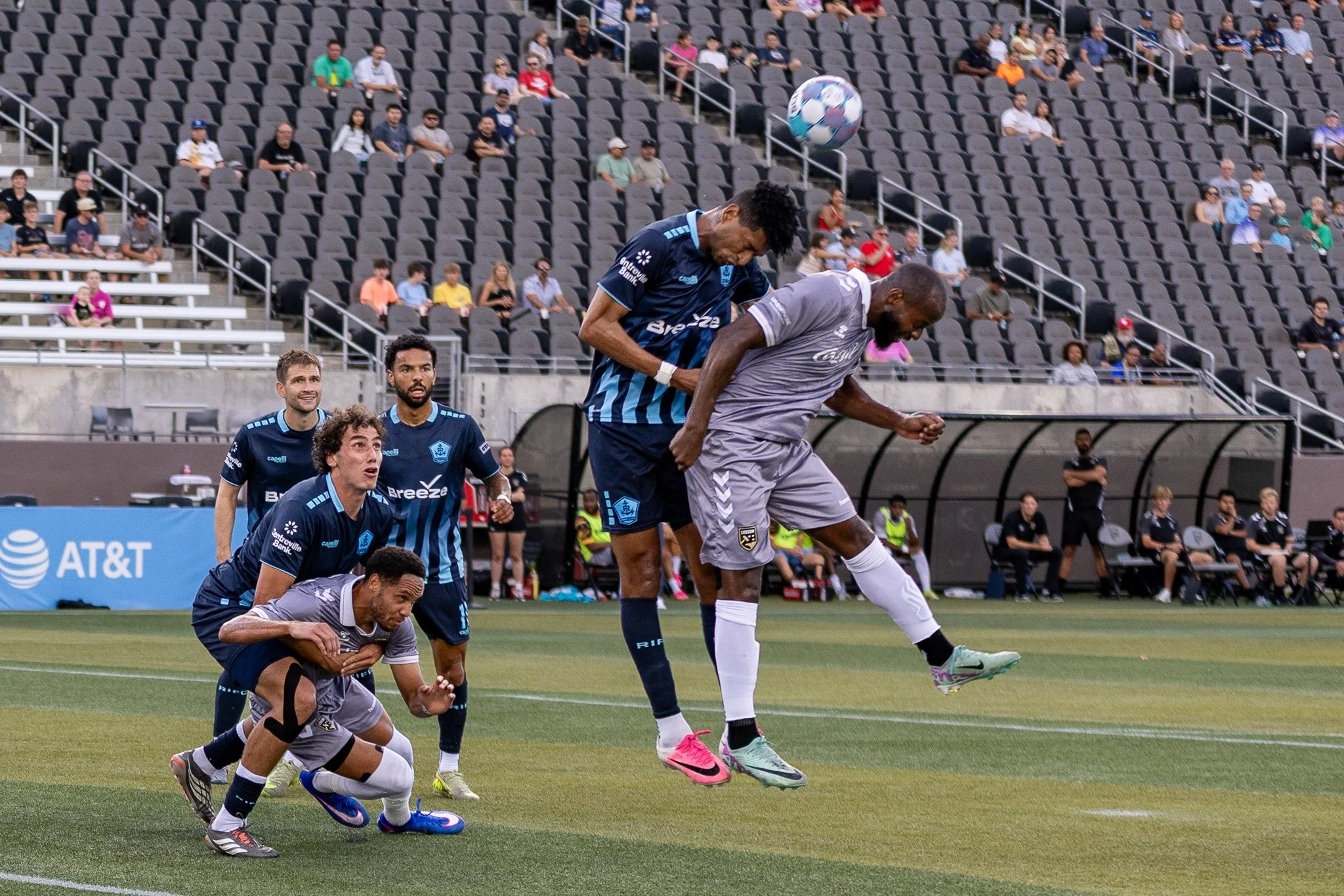 Soccer players compete for a header during a match in a stadium with spectators in the stands.