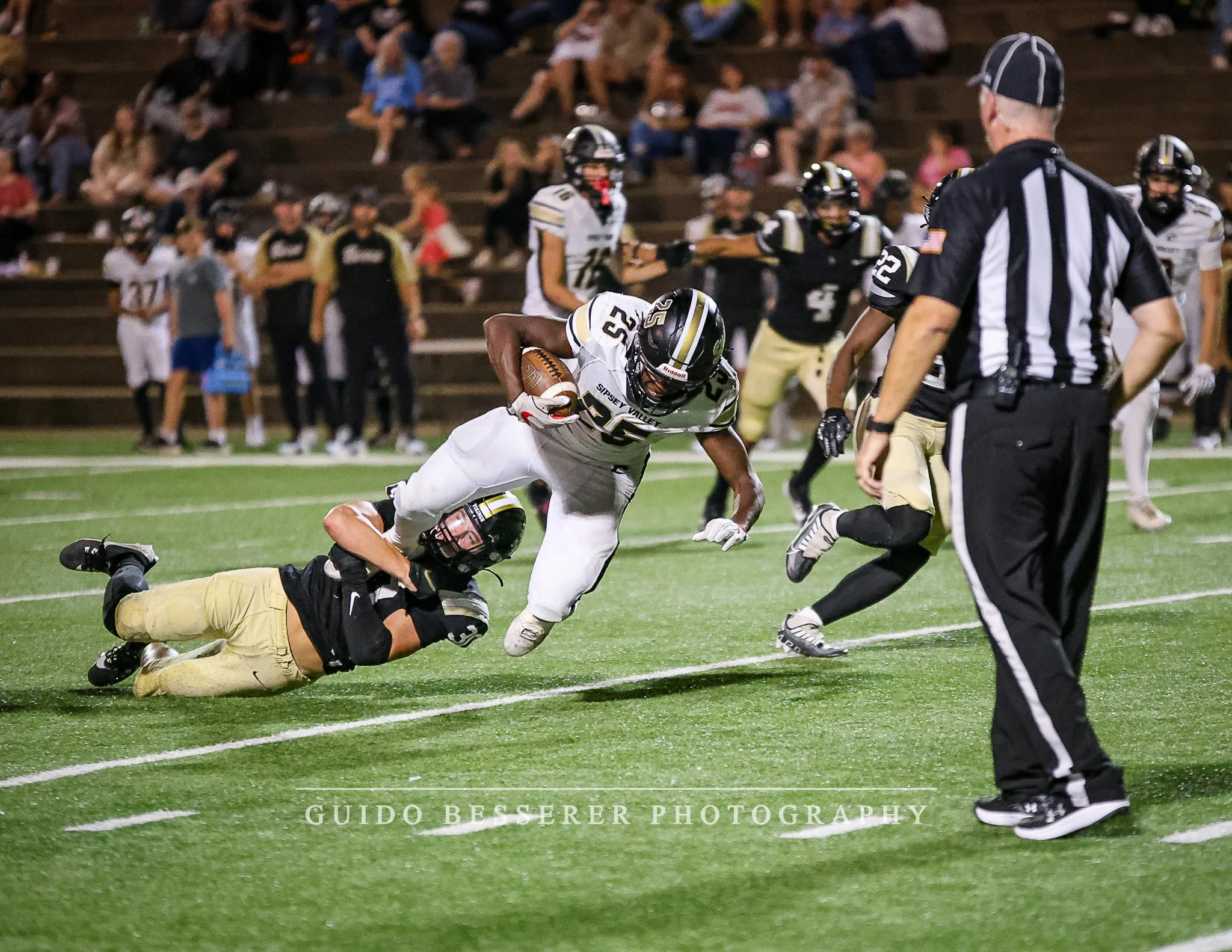 A football player in white uniform runs with the football, evading a tackle from a player in black and yellow uniform on a green field, with spectators watching from the bleachers in the background.