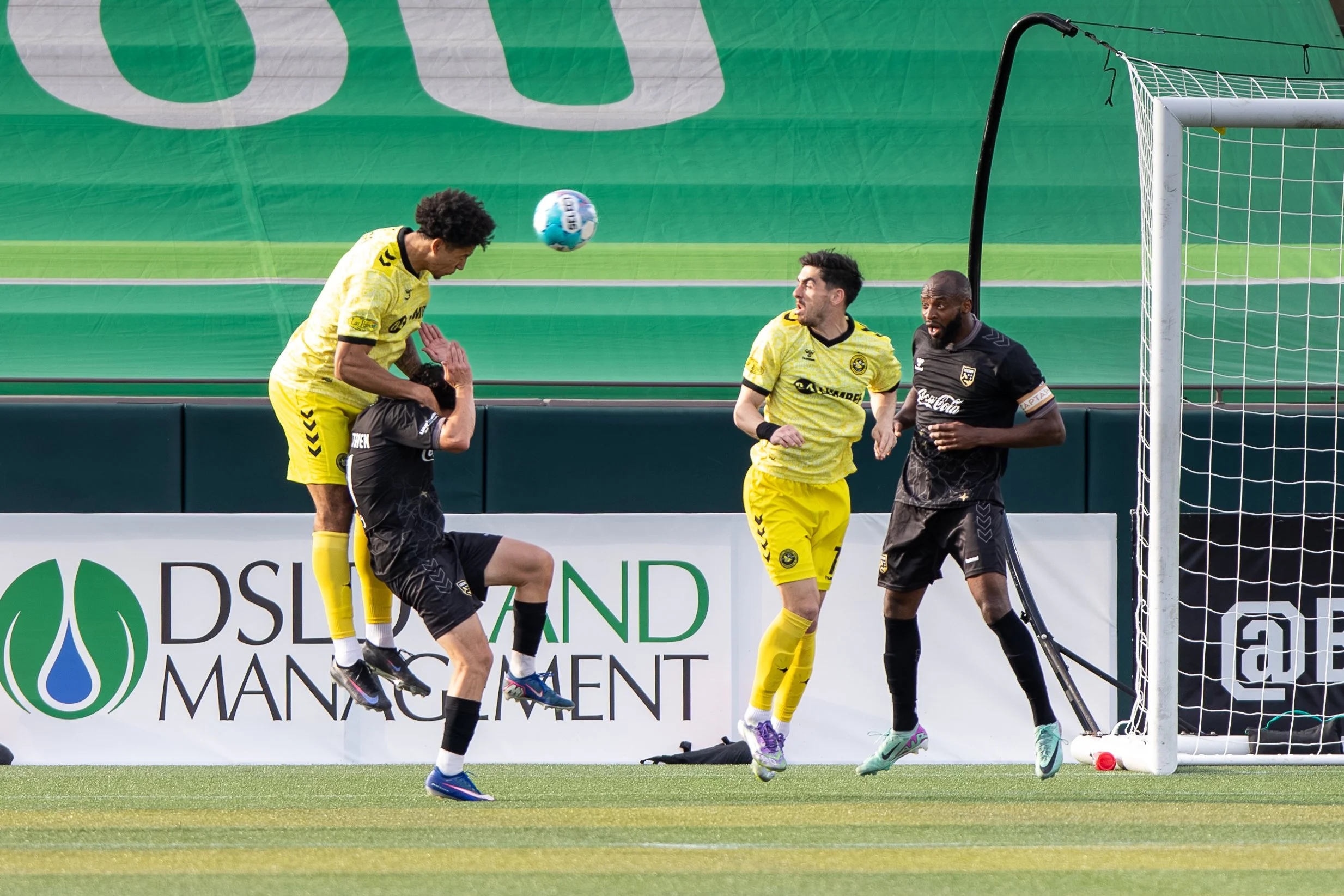 A soccer player in black being lifted by a teammate in yellow, with two more players in yellow observing, as they all look at the ball near the goalpost during a match.