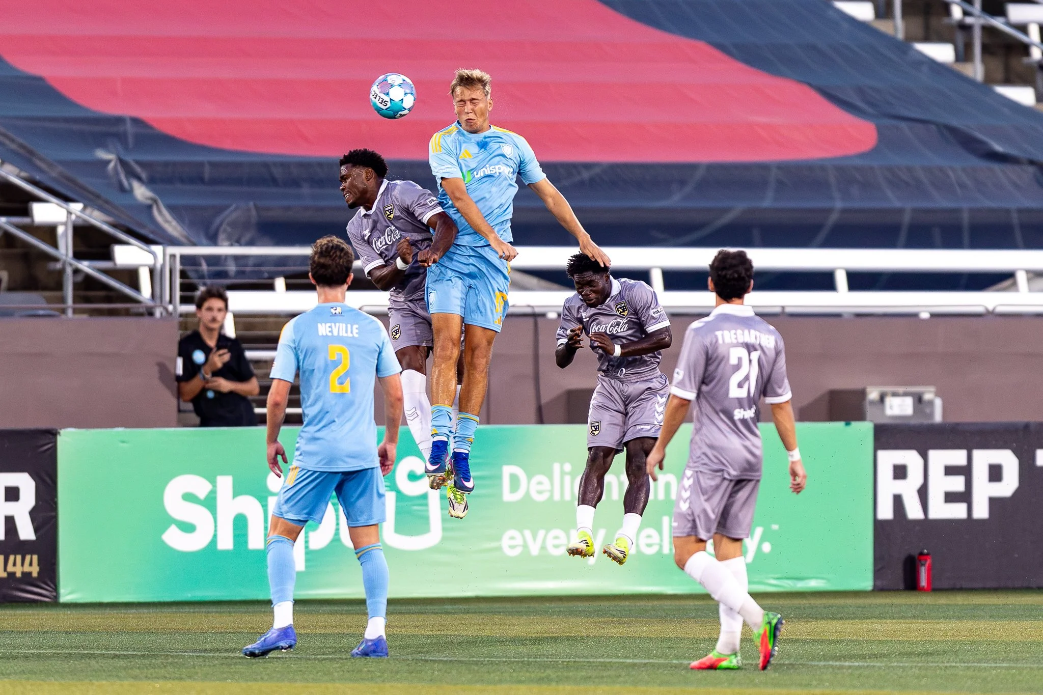 Soccer players during a match, with one player in a blue uniform jumping to head the ball while others in gray uniforms are nearby on the field.
