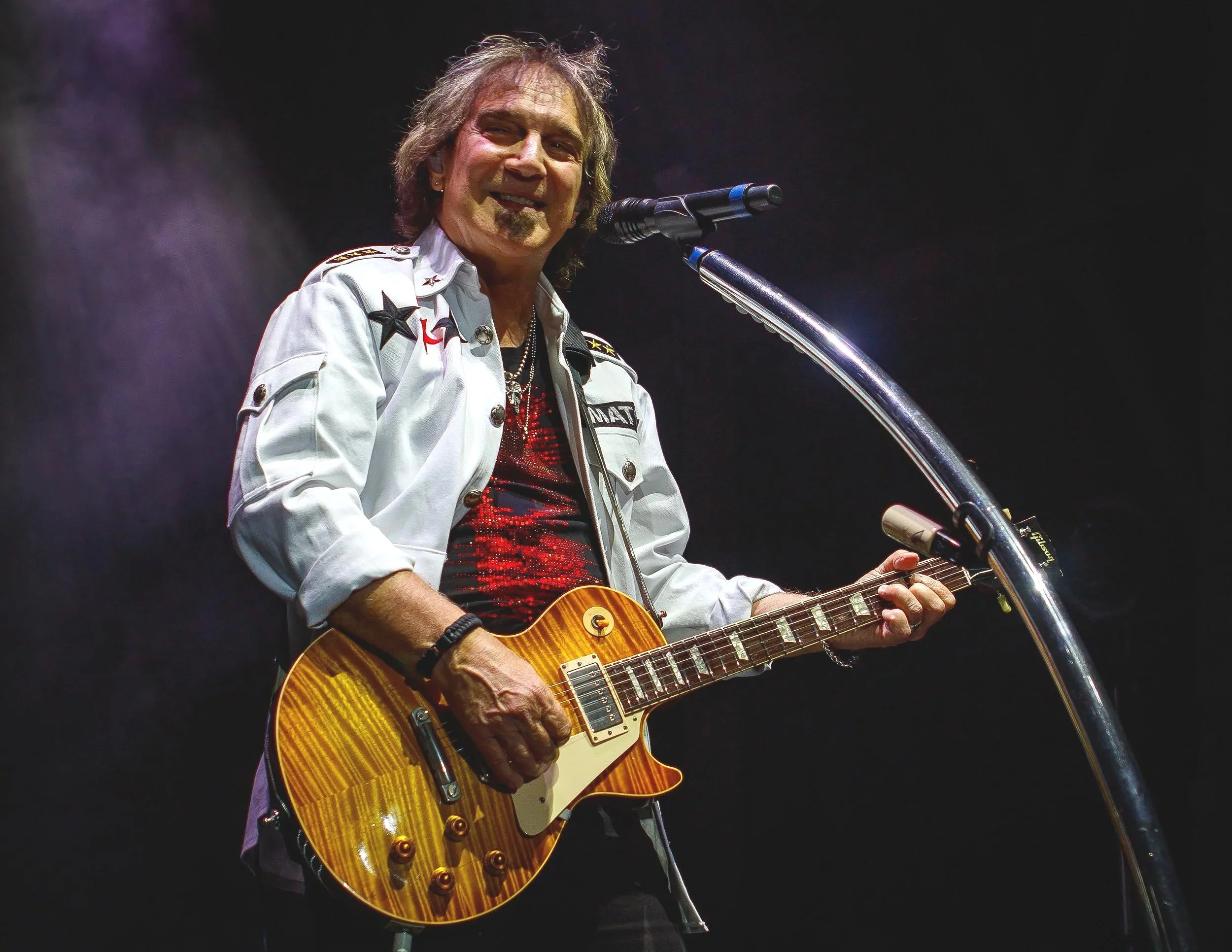 Dave Amato of REO Speedwagon performing on stage, playing an electric guitar, dressed in a white jacket with patches and a red sequined shirt, in front of a microphone with a dark background.