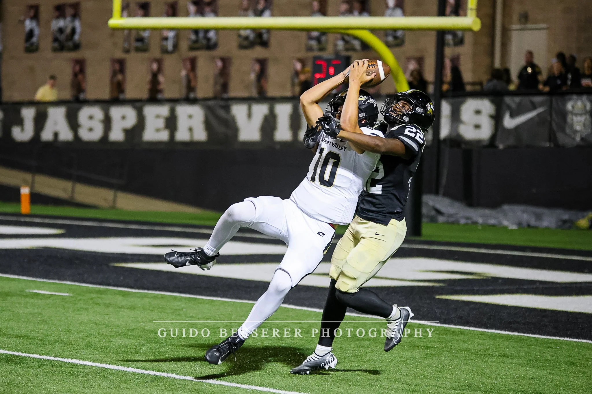 Two football players in black and white uniforms compete for the ball near the end zone, with one player jumping to catch it while the other tries to block. The game is taking place in a stadium.