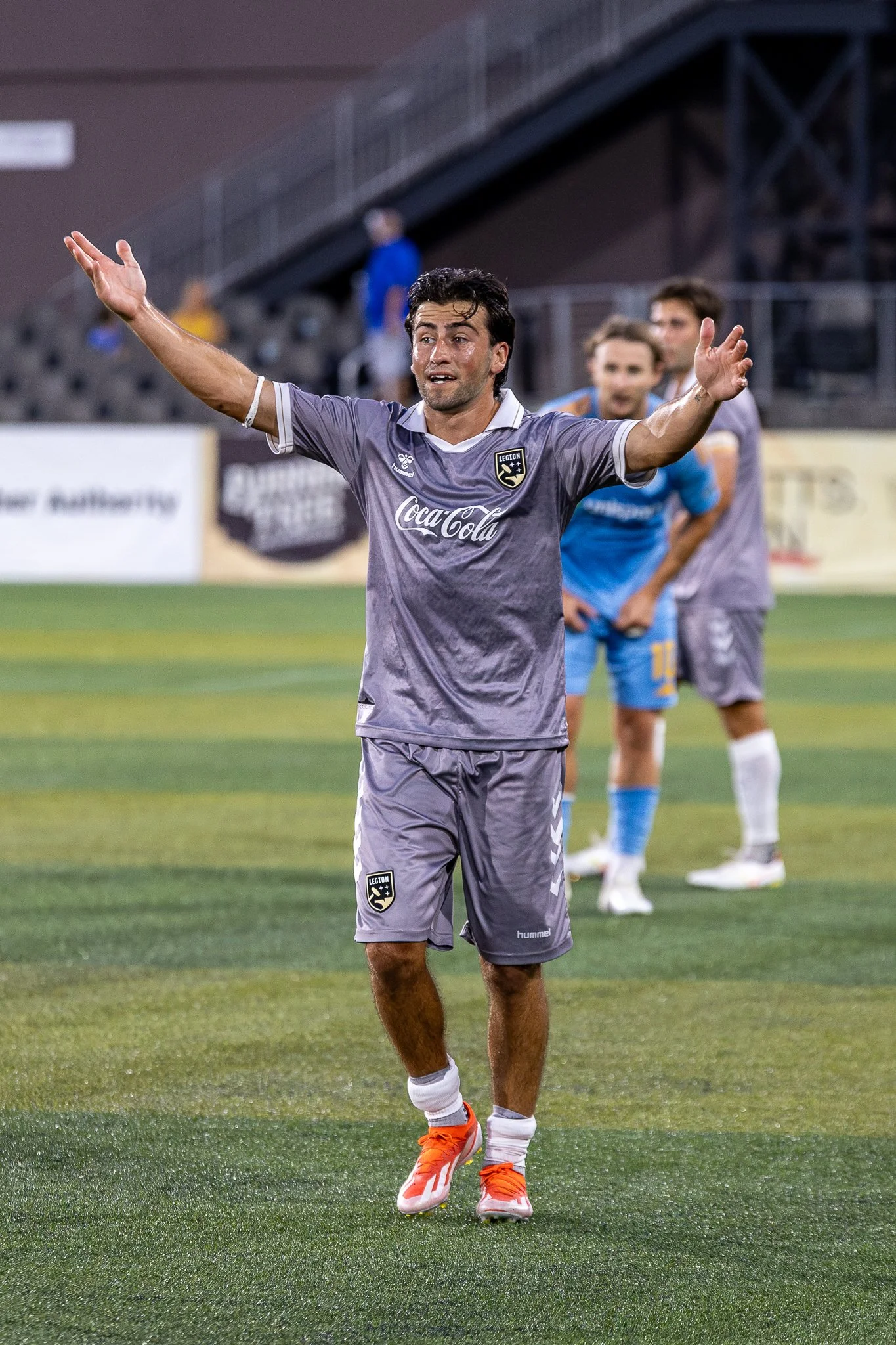 A male soccer player in a gray uniform with the logo 'LEGION' on the chest and shorts, celebrating on the field with arms outstretched. In the background, teammates and a stadium with empty seats are visible.