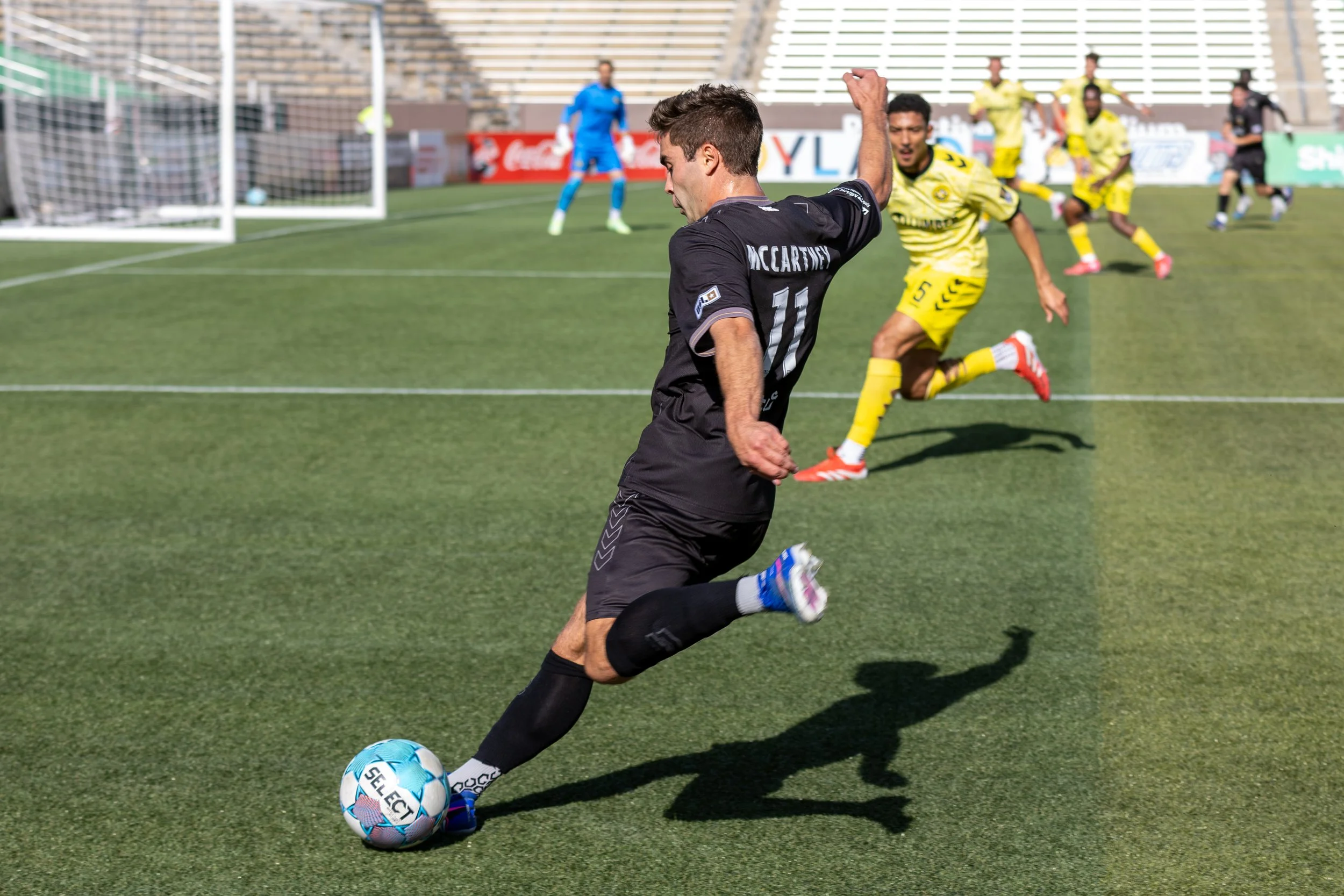 A soccer player in a black jersey with the number 11 is kicking a blue and white soccer ball on a field, with other players and a goalkeeper in yellow and black jerseys in the background.