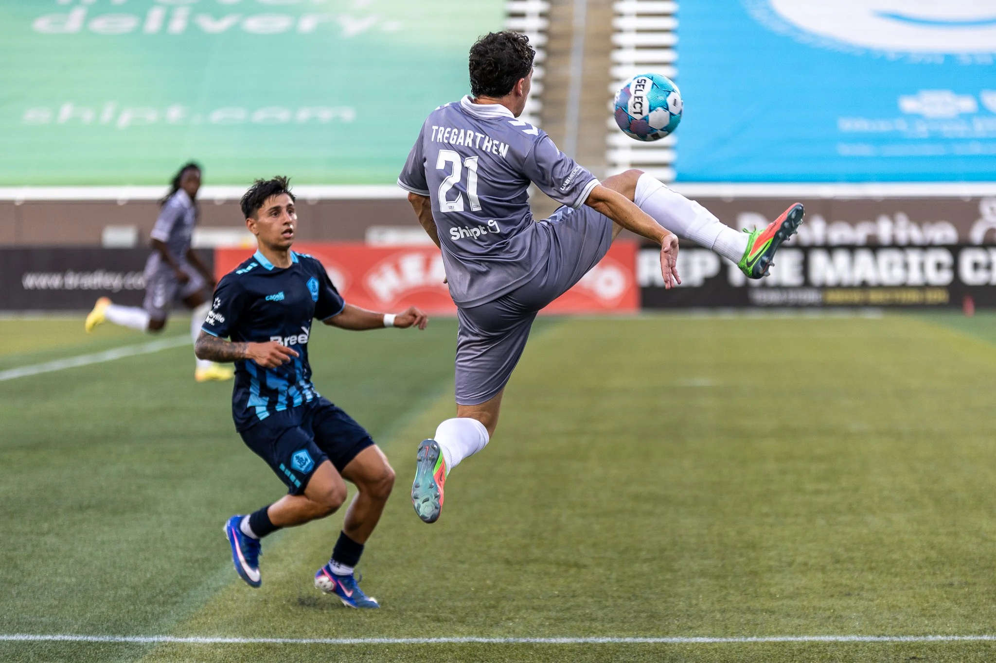 A soccer player wearing gray jersey number 21 jumps to kick a blue and white soccer ball during a match. Two other players are visible in the background on the field.