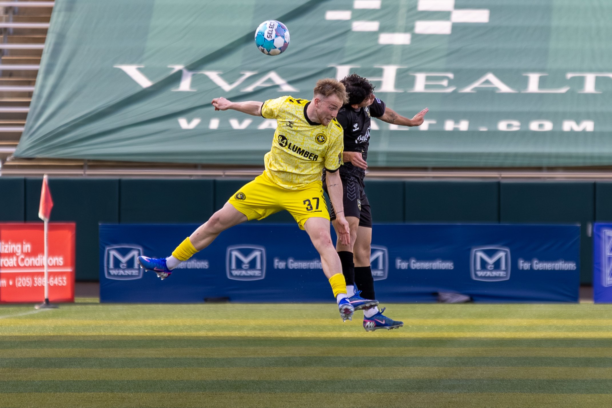 Two soccer players compete for the ball near the corner flag during a match. One player, wearing a yellow jersey and shorts, is lunging forward, while the other, dressed in black, is closely contesting.