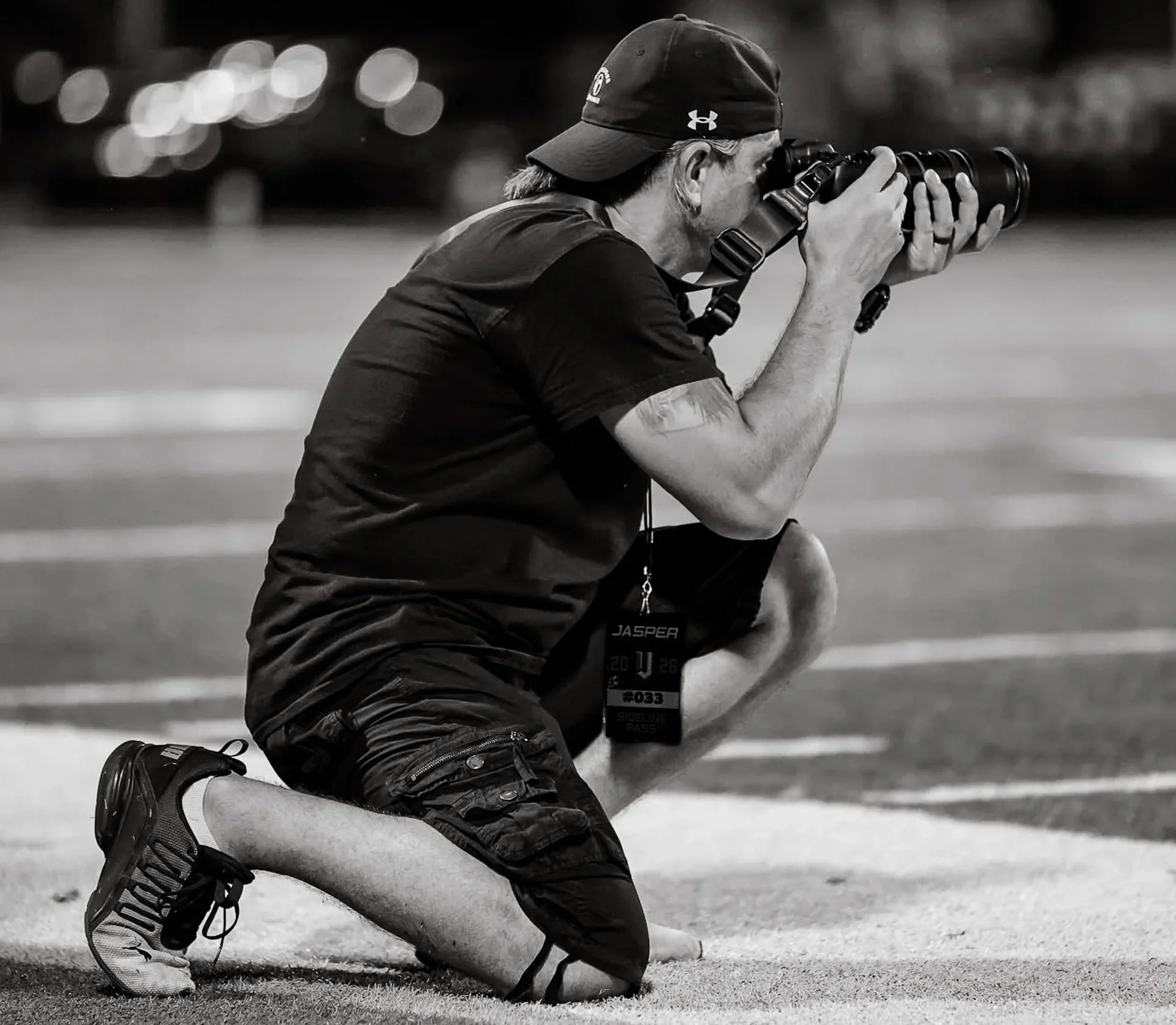 A person with long hair wearing a cap and athletic clothing kneeling on a field, taking photographs with a camera with a long telephoto lens.