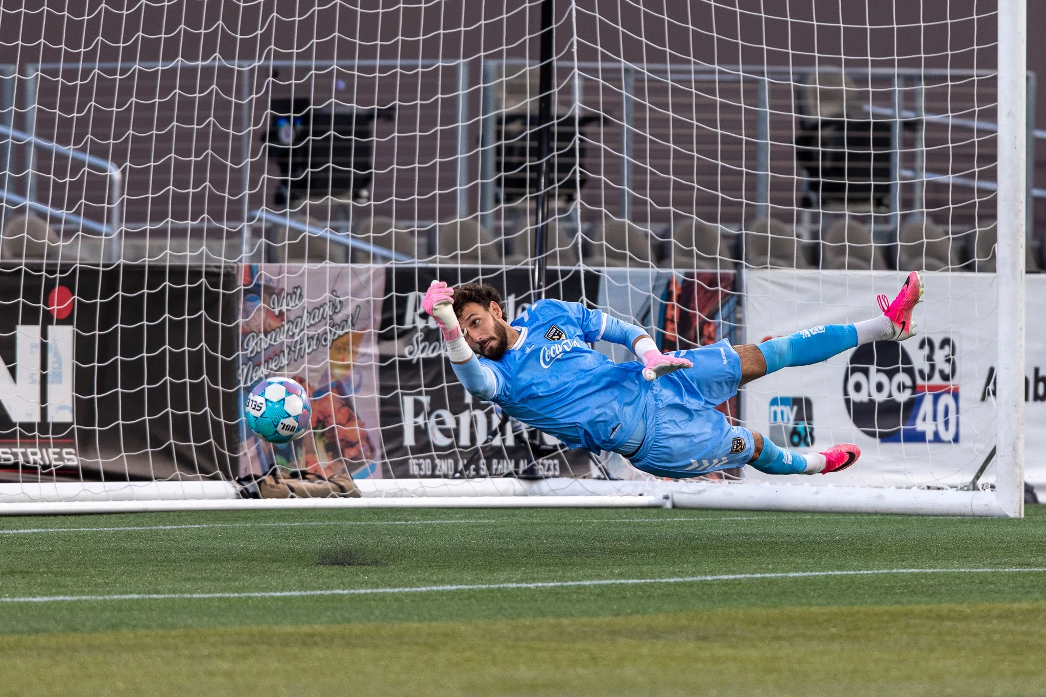 A soccer goalkeeper in a blue uniform dives to their left attempting to save a ball heading into the net, with a goalpost and net behind them.