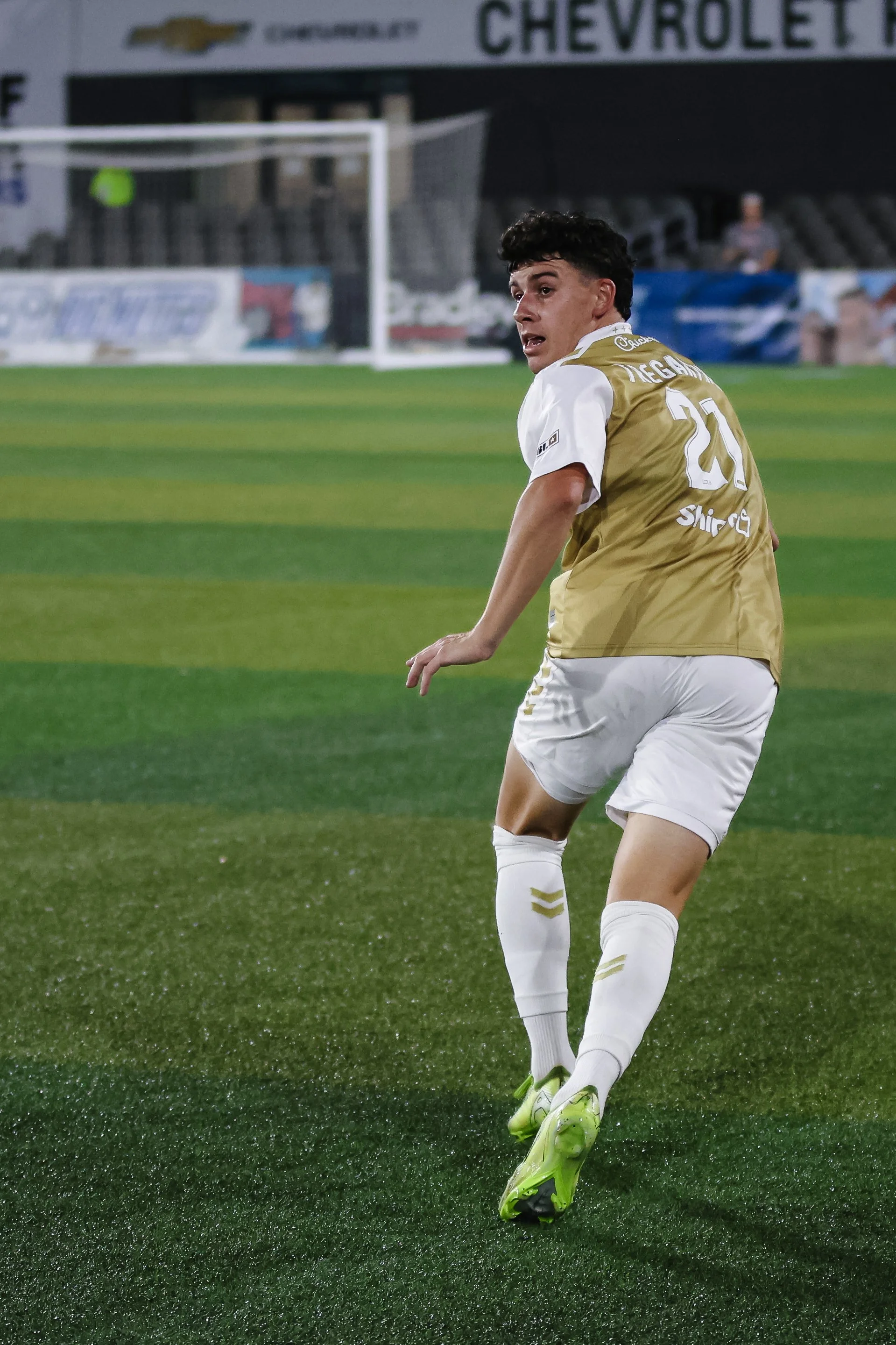 A soccer player wearing a gold and white uniform on the field, facing away, with a stadium in the background.