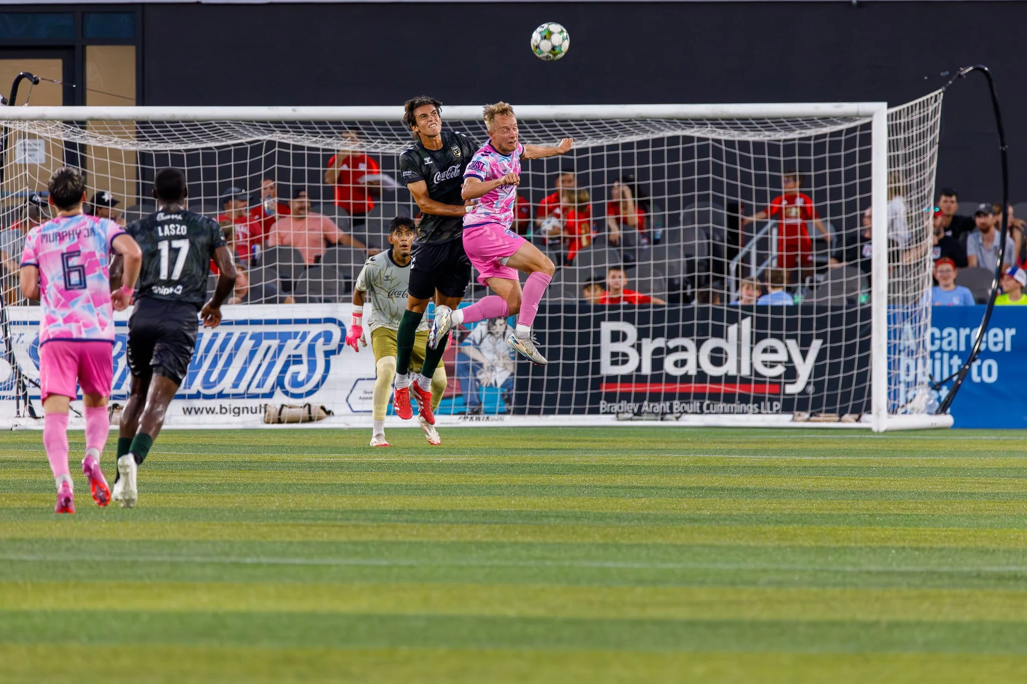 Soccer players jumping to head the ball during a match, with spectators watching in the background.