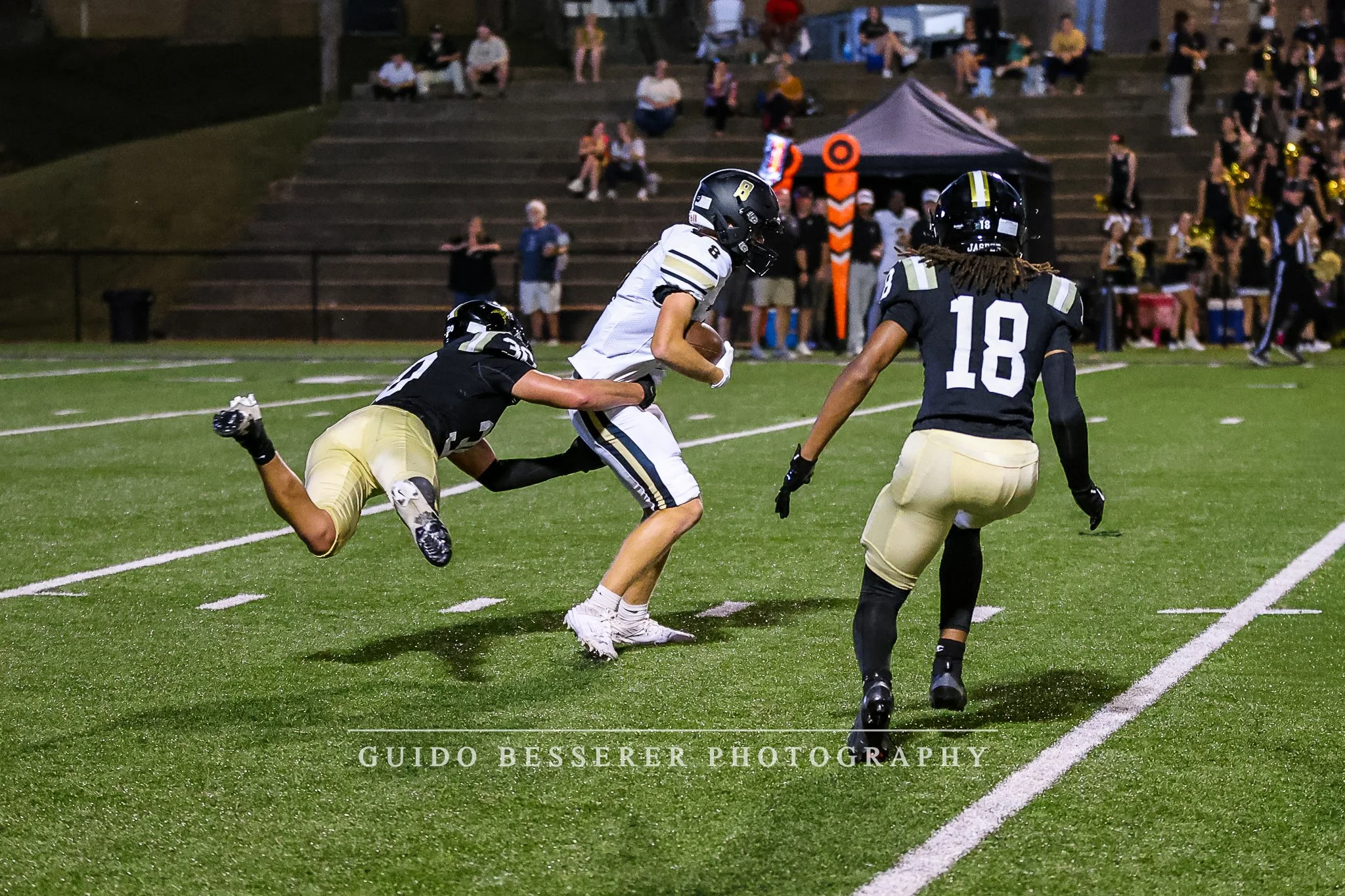 A football player in a white jersey running with the ball while two opposing players in black jerseys try to tackle him during a game on a football field at night.