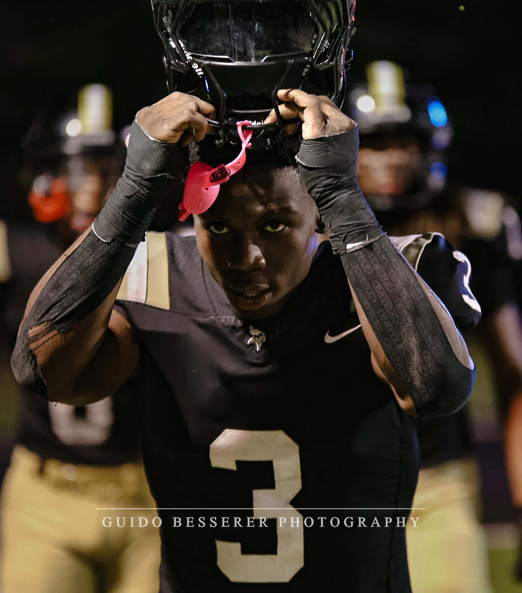 A football player in a black jersey with the number 3 puts on a helmet at night.