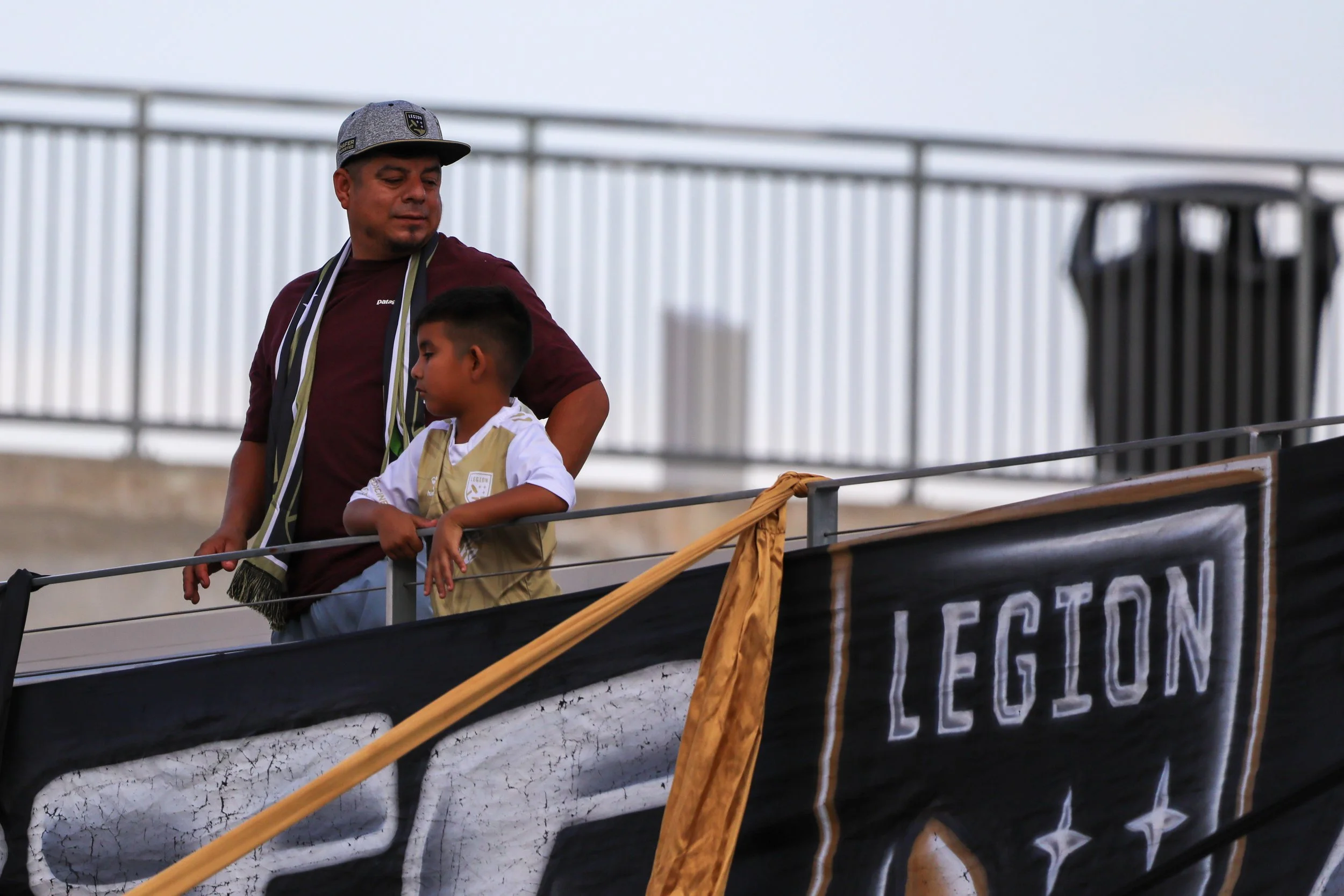 A man and a young boy standing on a balcony, with the man looking at the boy. The man wears a gray cap and a maroon shirt, while the boy wears a white shirt and a beige vest. There is a banner with the word 'LEGION' hanging on the balcony railing.