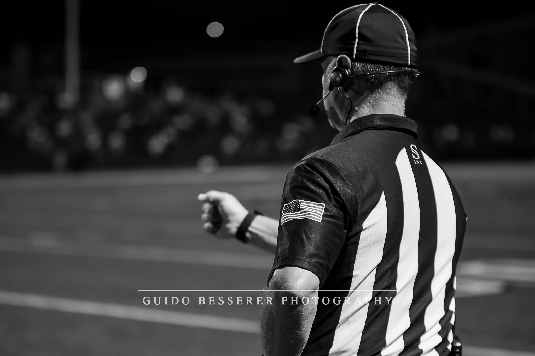 A black and white photo of a football referee wearing a US national team jersey, a cap, and a headset, standing on the field during a night game.