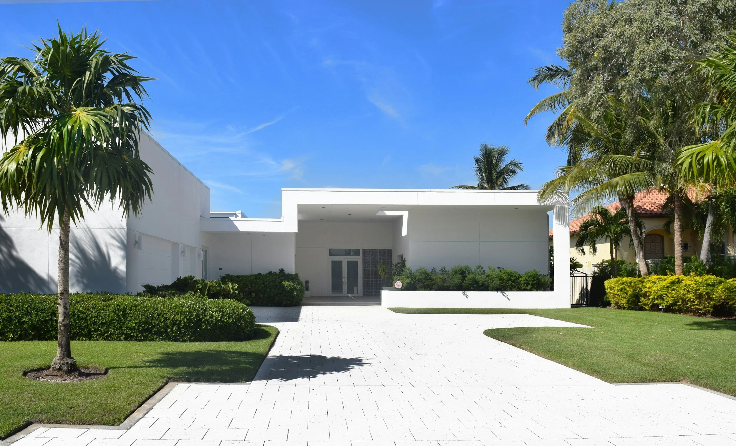Modern white residential building with a minimalist design, surrounded by green bushes and palm trees, under a clear blue sky.