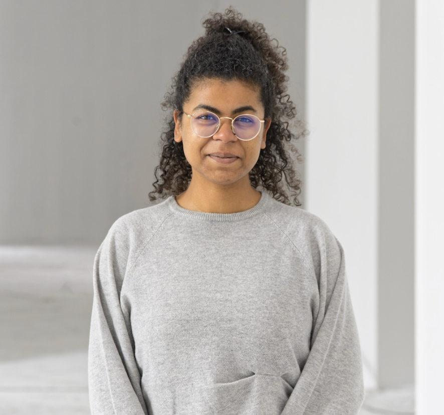 Young woman with curly hair and glasses wearing a gray sweatshirt standing indoors.