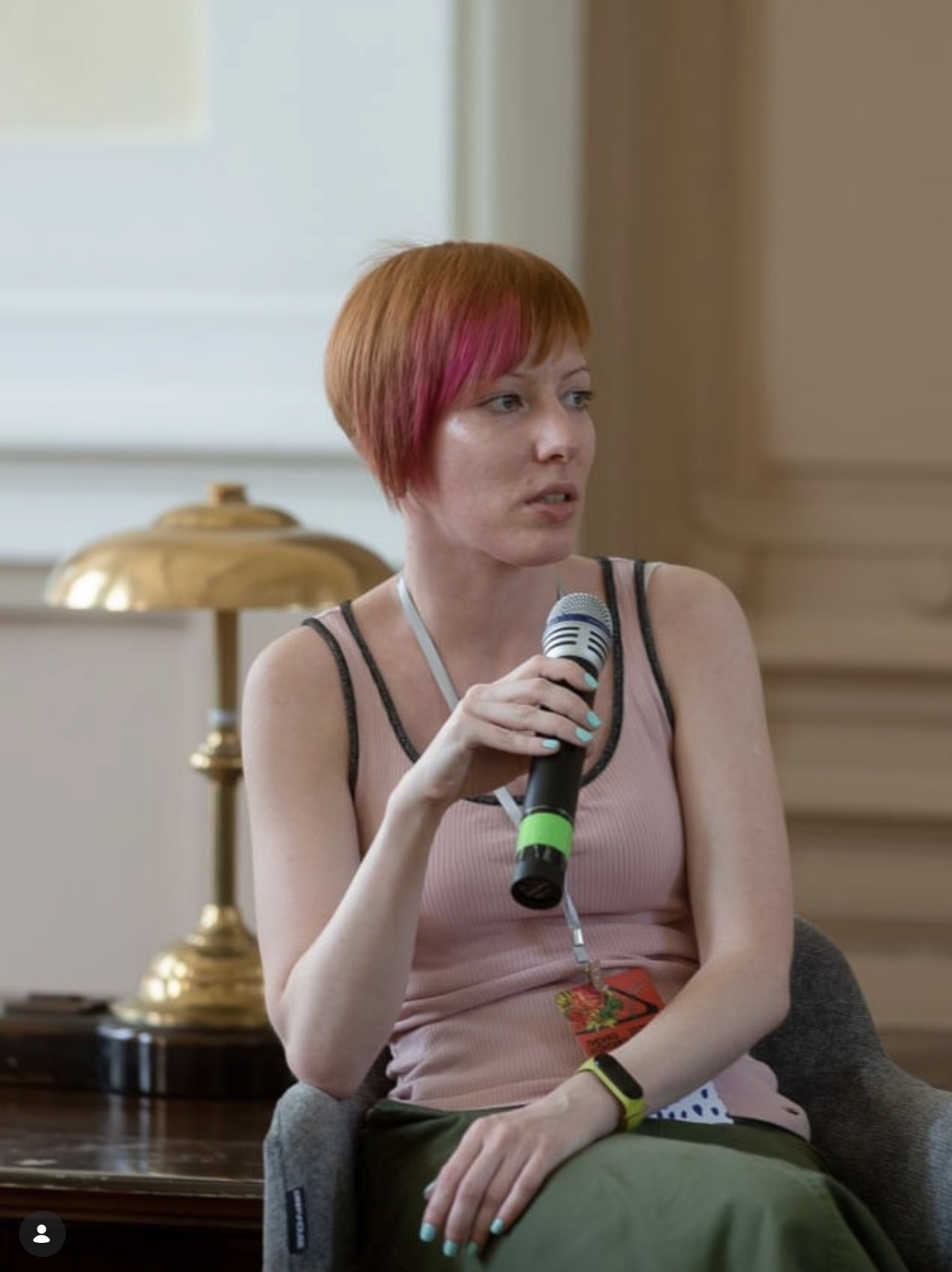 A woman with short red hair and pink highlights is speaking into a microphone during a panel or presentation. She is wearing a pink sleeveless top, green pants, and a smartwatch. She is seated in a room with beige walls and a gold lamp behind her.