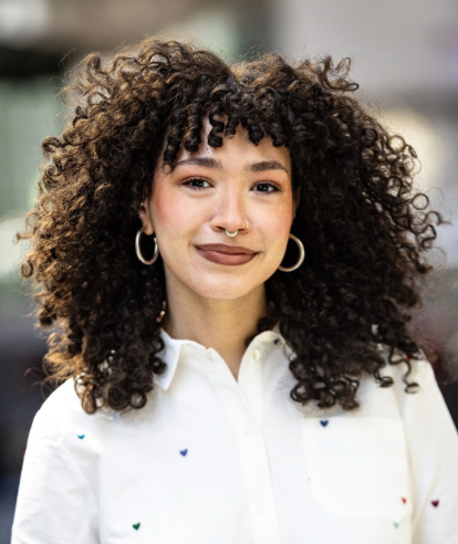 Young woman with curly hair, nose ring, hoop earrings, and wearing a white shirt with small colorful hearts, smiling indoors.
