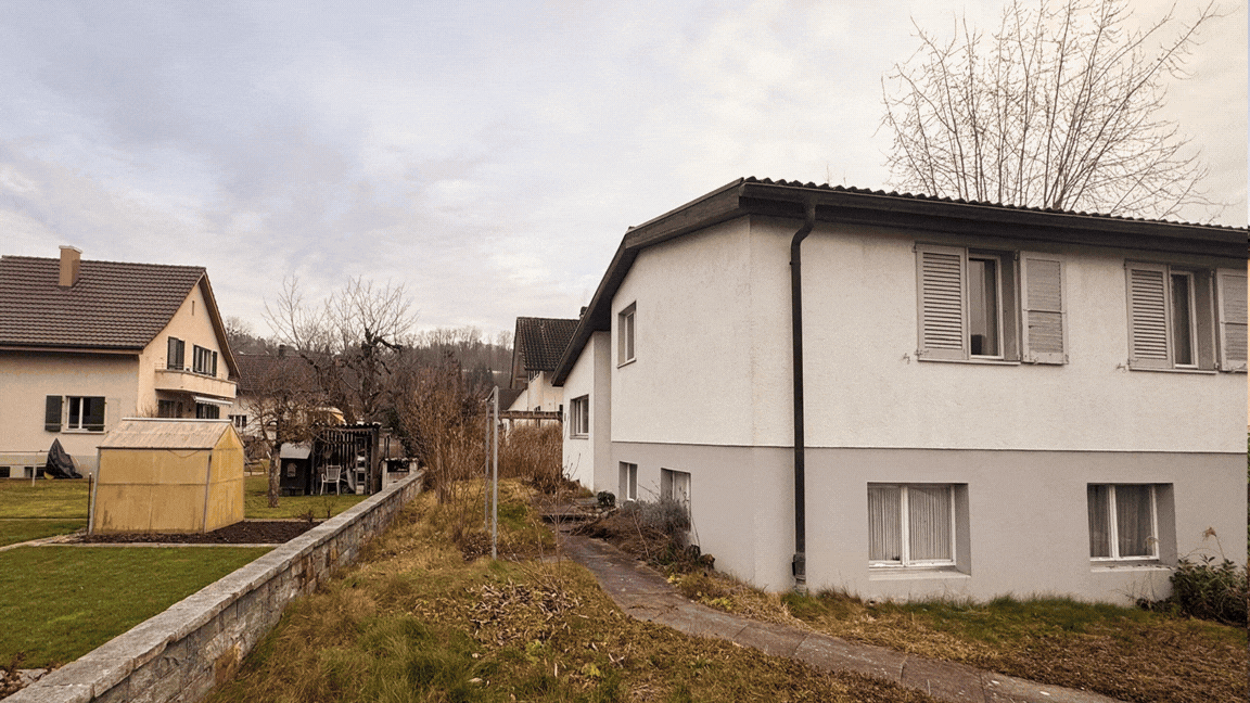 Stadthaus mit weißen Wänden und mehreren Fenstern, Garten mit Rasen, Zaun und Gartenhaus, im Hintergrund weitere Häuser und Bäume, bewölkter Himmel.