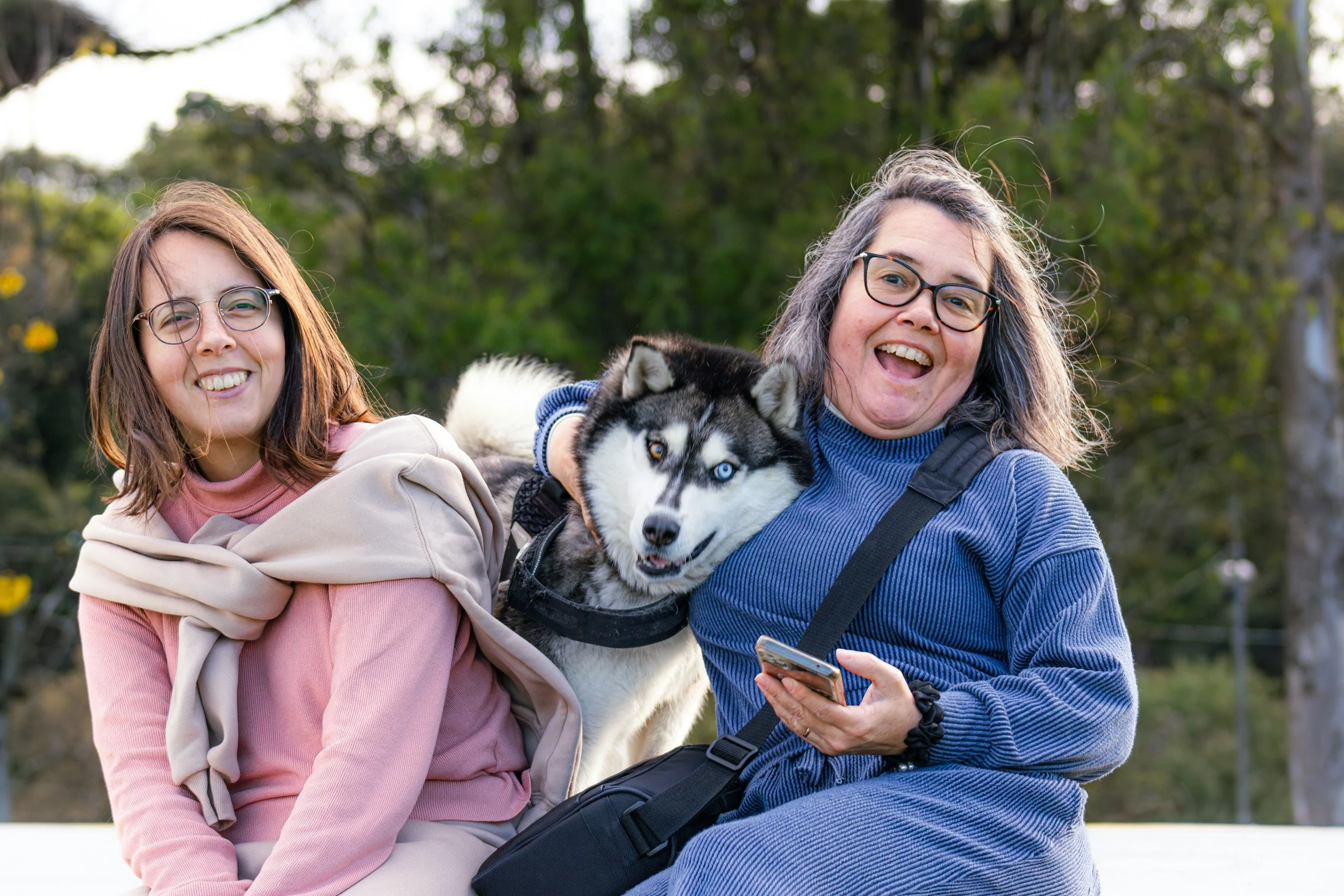 Two women sitting outdoors with a Siberian Husky dog between them, smiling and enjoying the day.