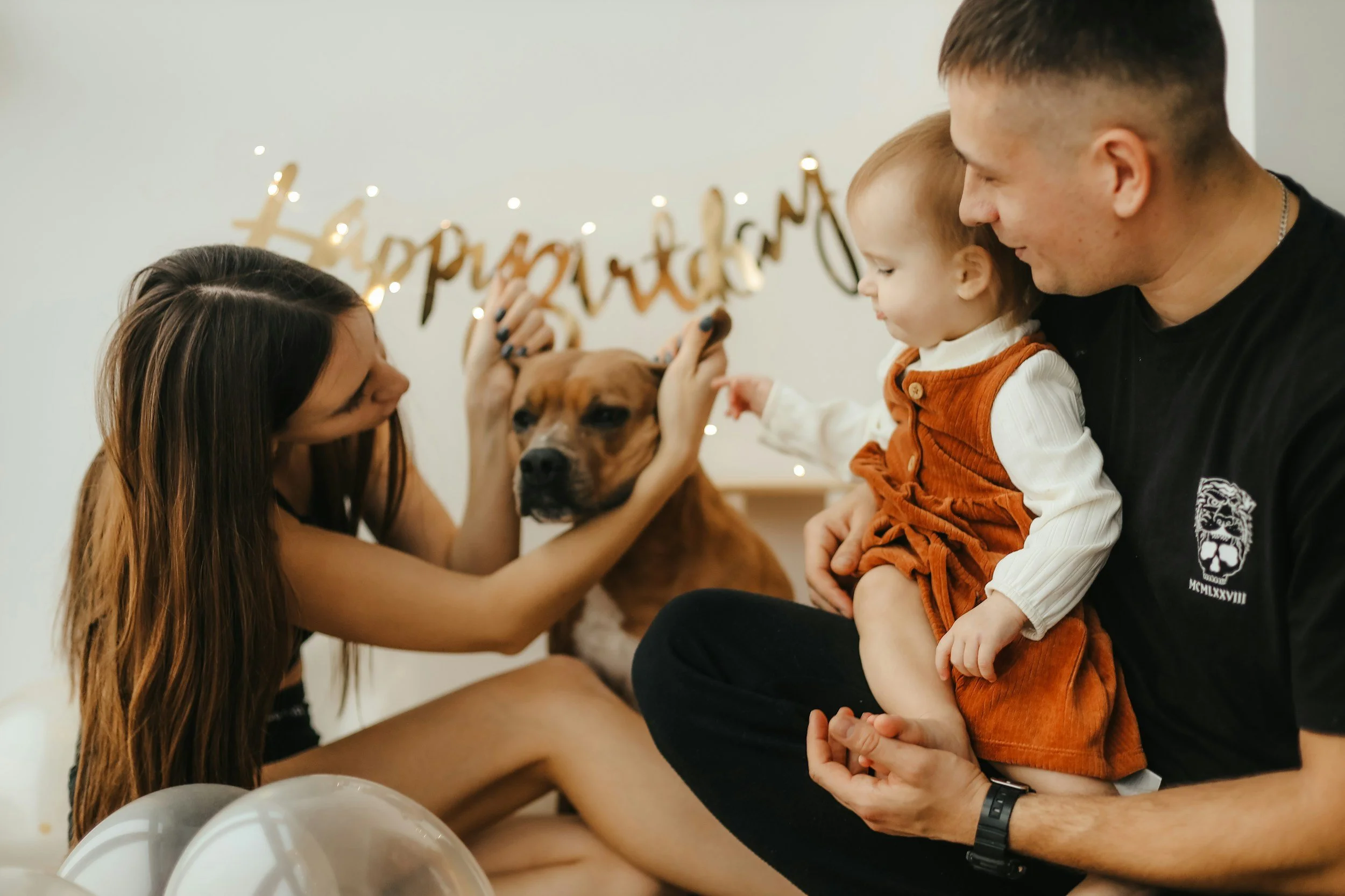 A family celebrating a birthday with a woman, a man, a young girl, and a dog. The woman is petting the dog while the man holds the young girl, who is pointing at the dog. There are balloons in the foreground and a birthday banner with lights behind them.